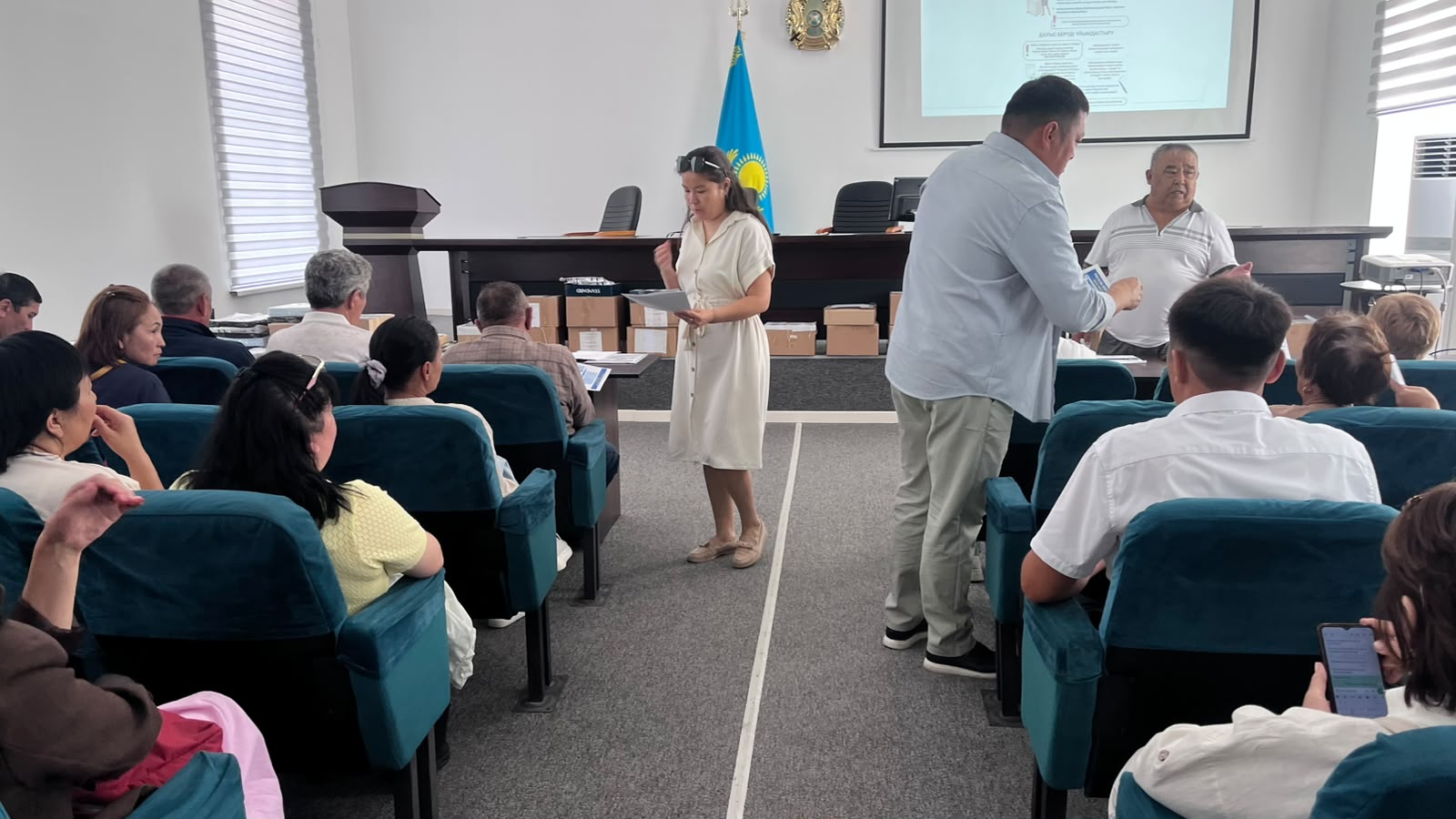 Жиналыс залында баяндама жасап тұрған қыз және тыңдаушылар.