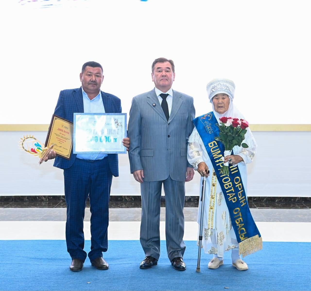 Диплом мен гүл ұстаған екі ер адам мен лента таққан қарт әйелдің салтанатты суреті.