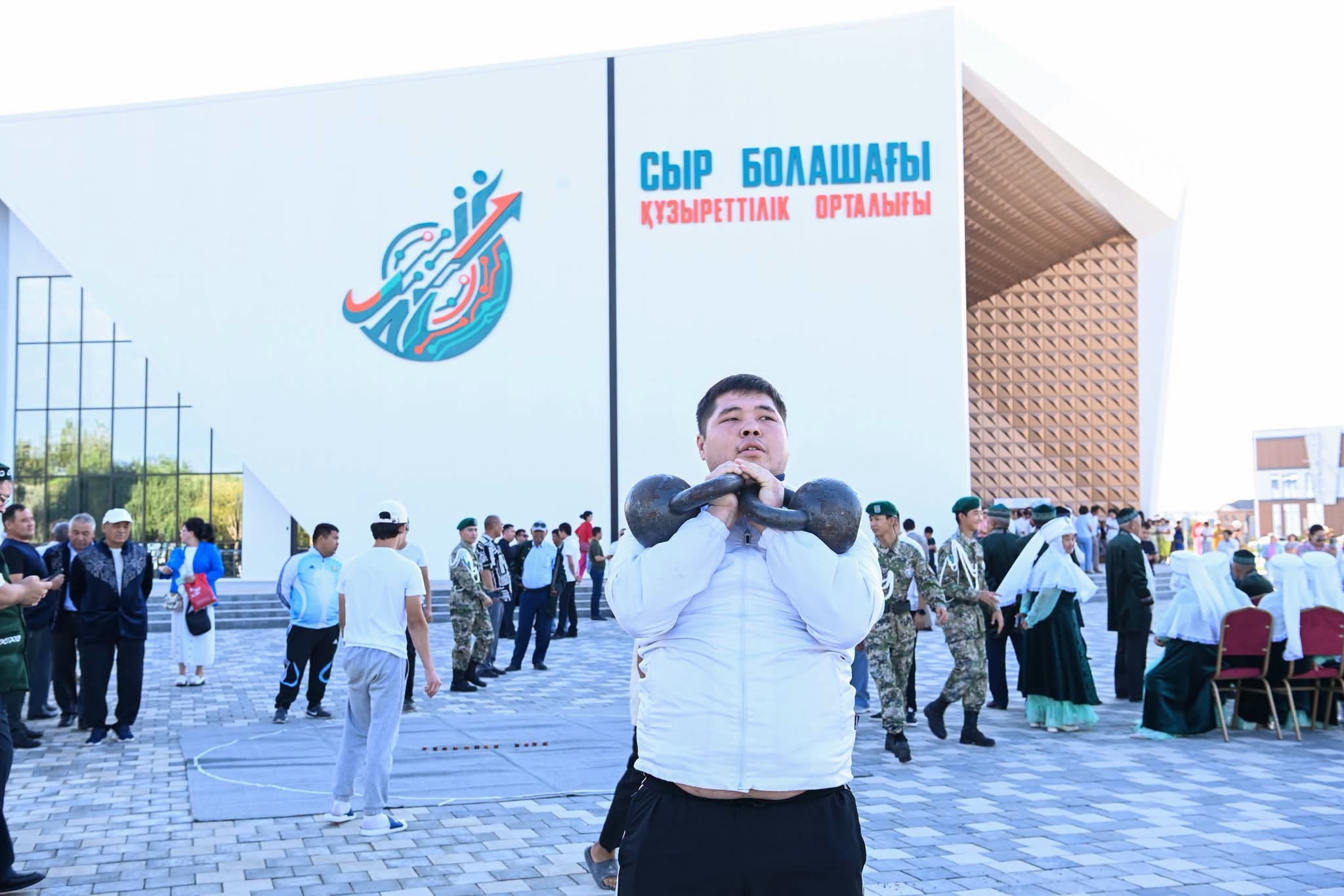 «Сыр болашағы» орталығының алдында гиря көтеріп жатқан спортшы және жиналған адамдар.