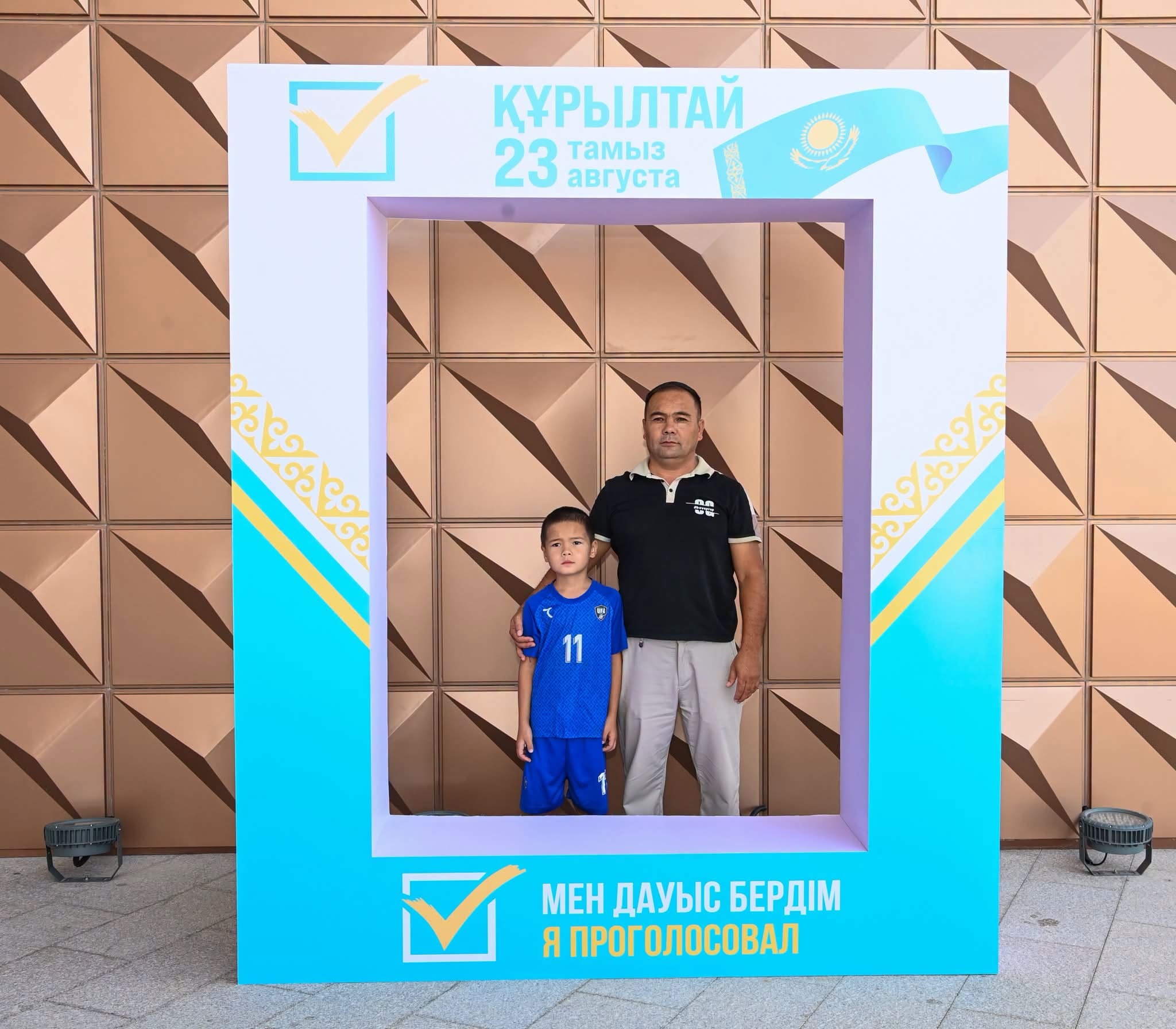 «Құрылтай 23 тамыз» жазуы бар фотозонада тұрған ер адам мен бала.