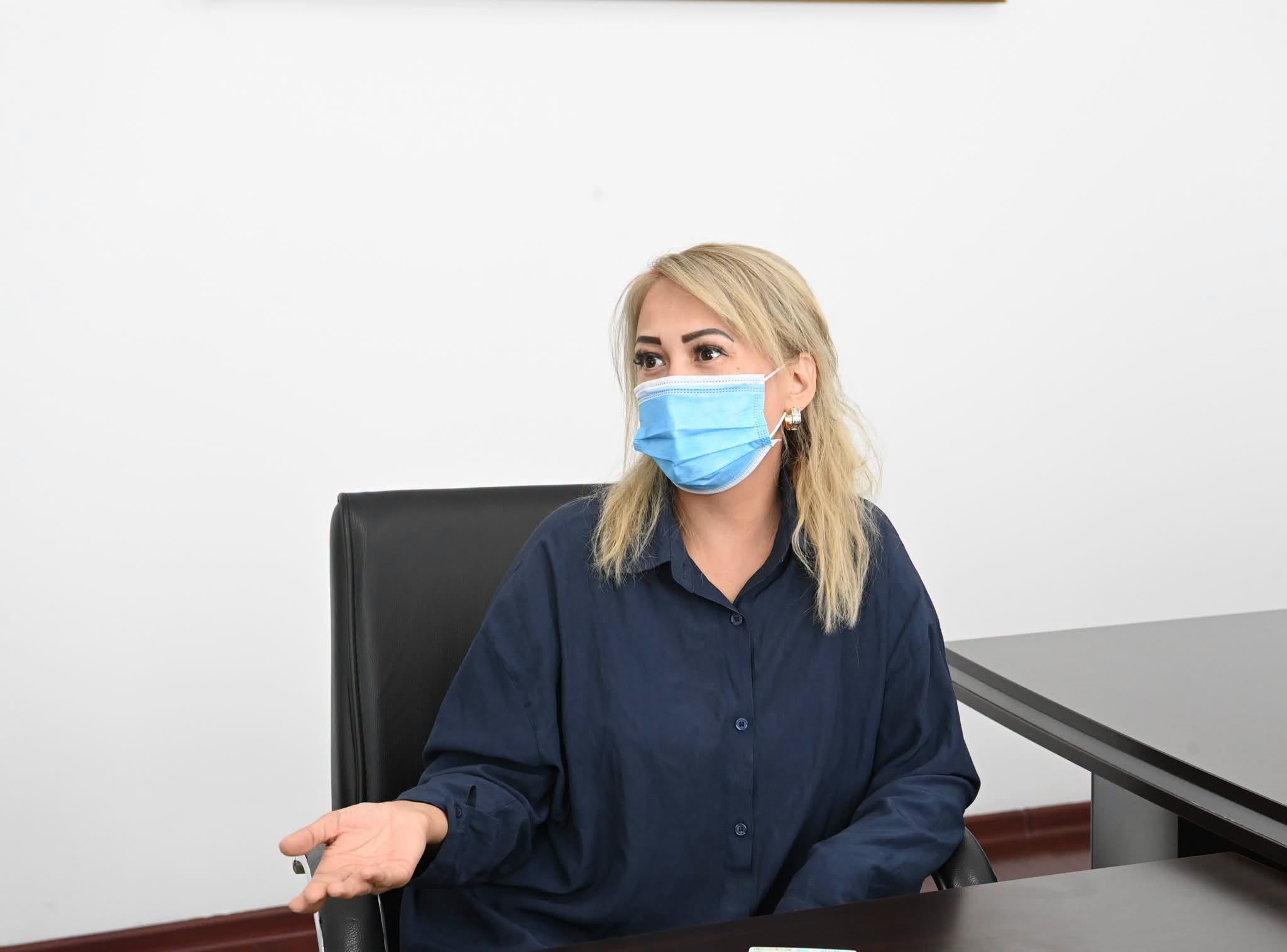 Көк жейде мен бетперде киген әйел адам келіссөз үстелінде отыр.