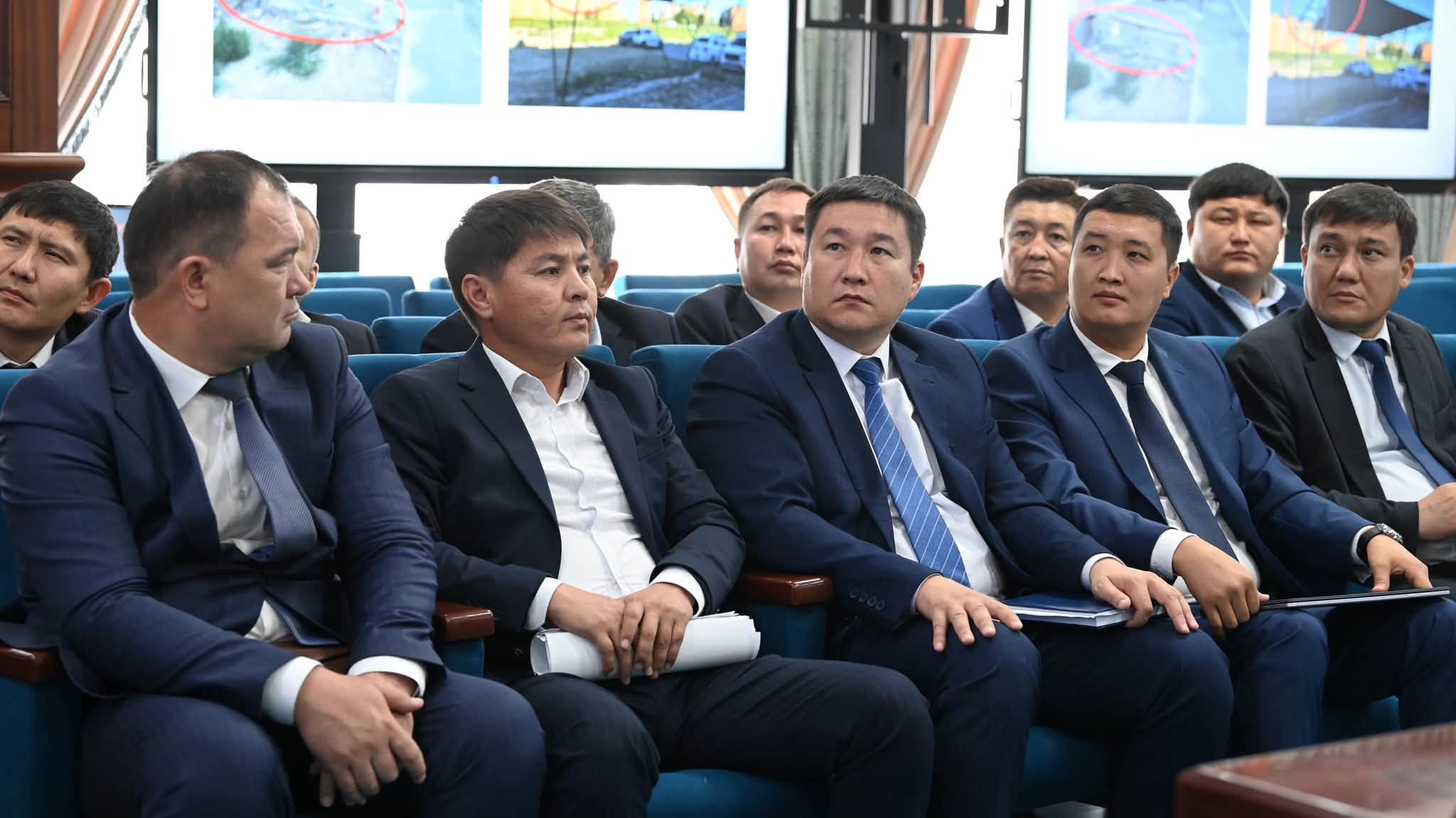 Көк костюм киген ер адамдар жиналыста отырып, экрандағы презентацияны тамашалап жатыр.