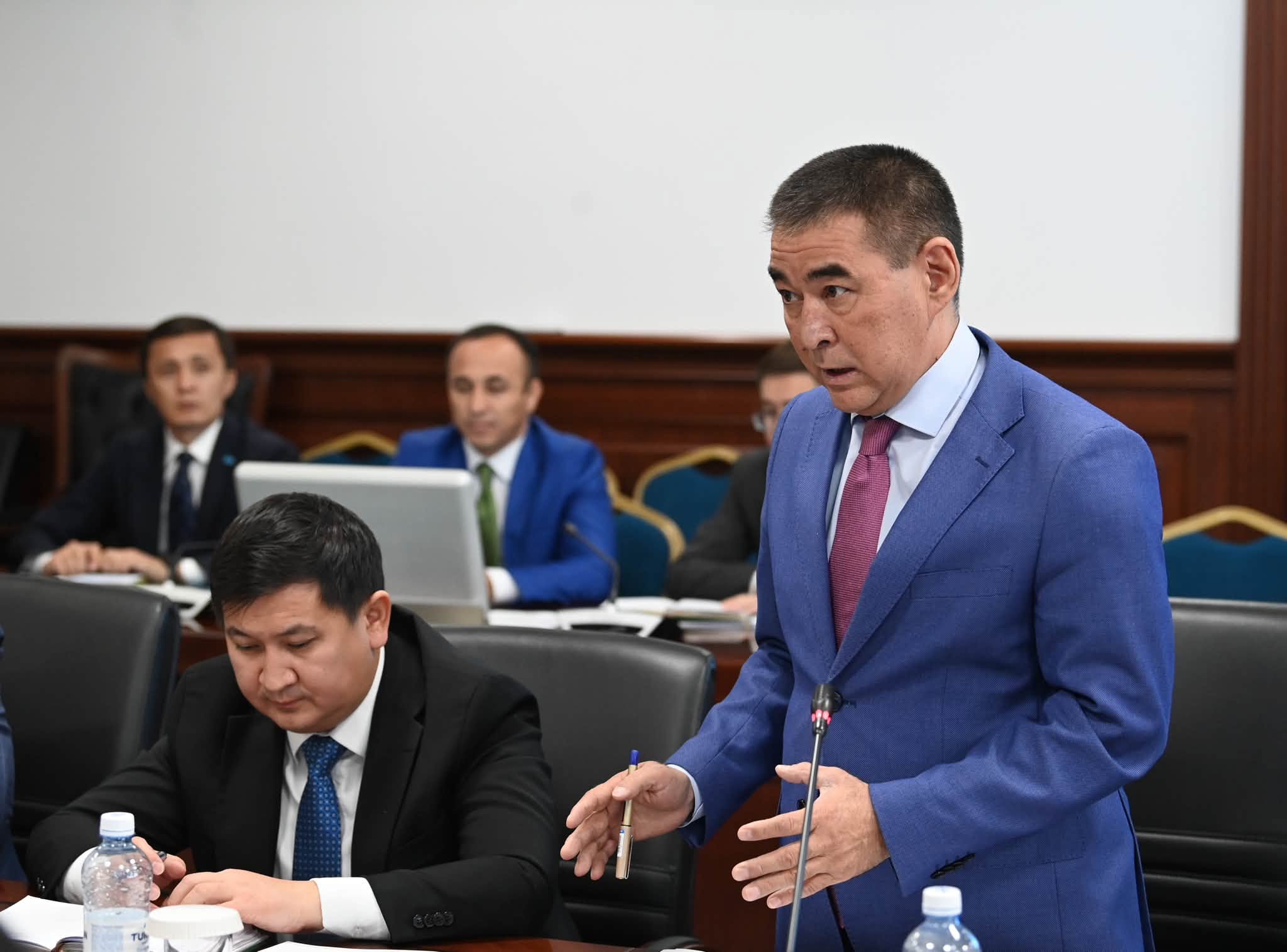 Көк костюм киген ер адам жиналыста сөйлеп тұр.