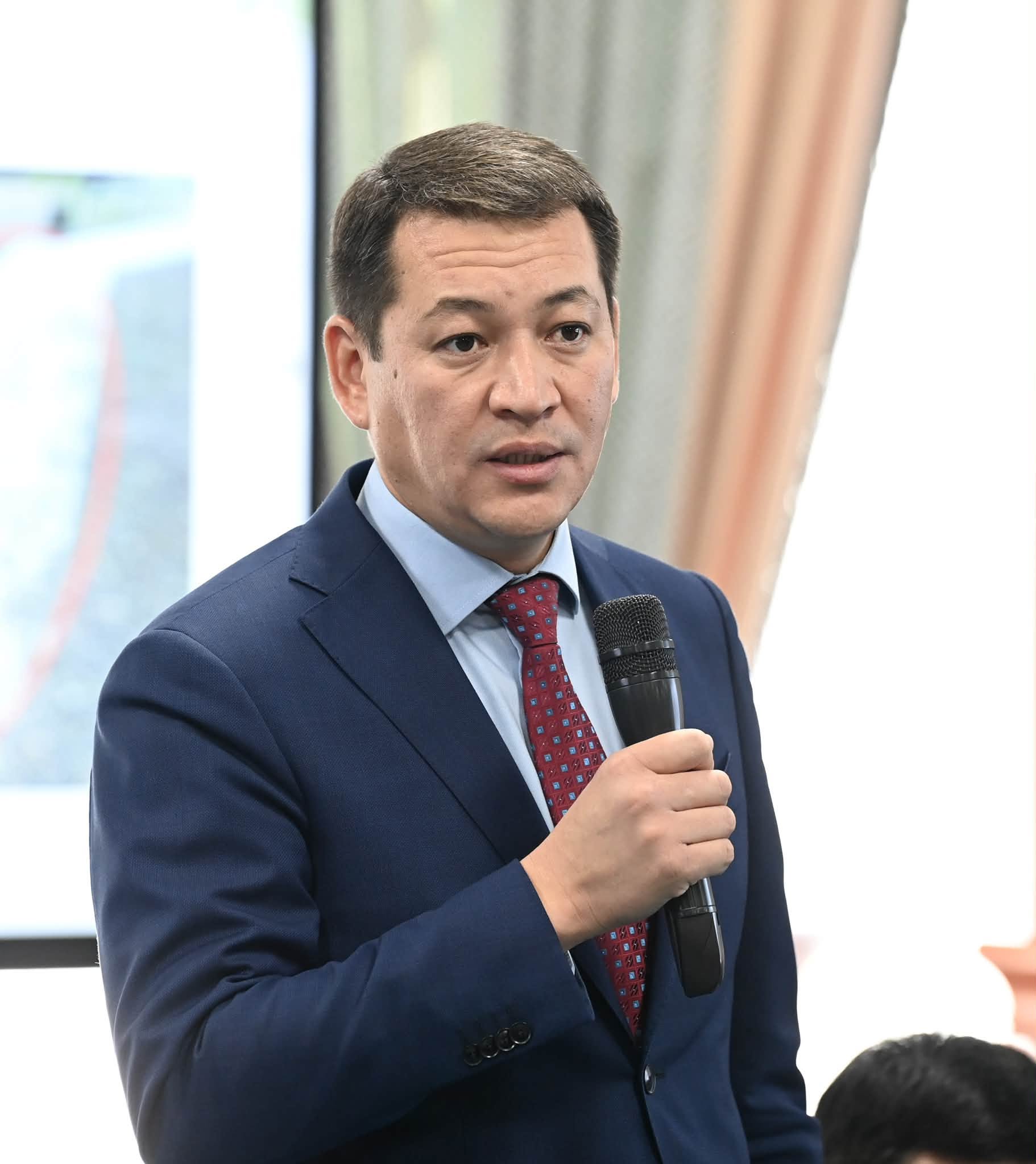 Көк костюм мен қызыл галстук киген, қолына микрофон ұстаған ер адам.