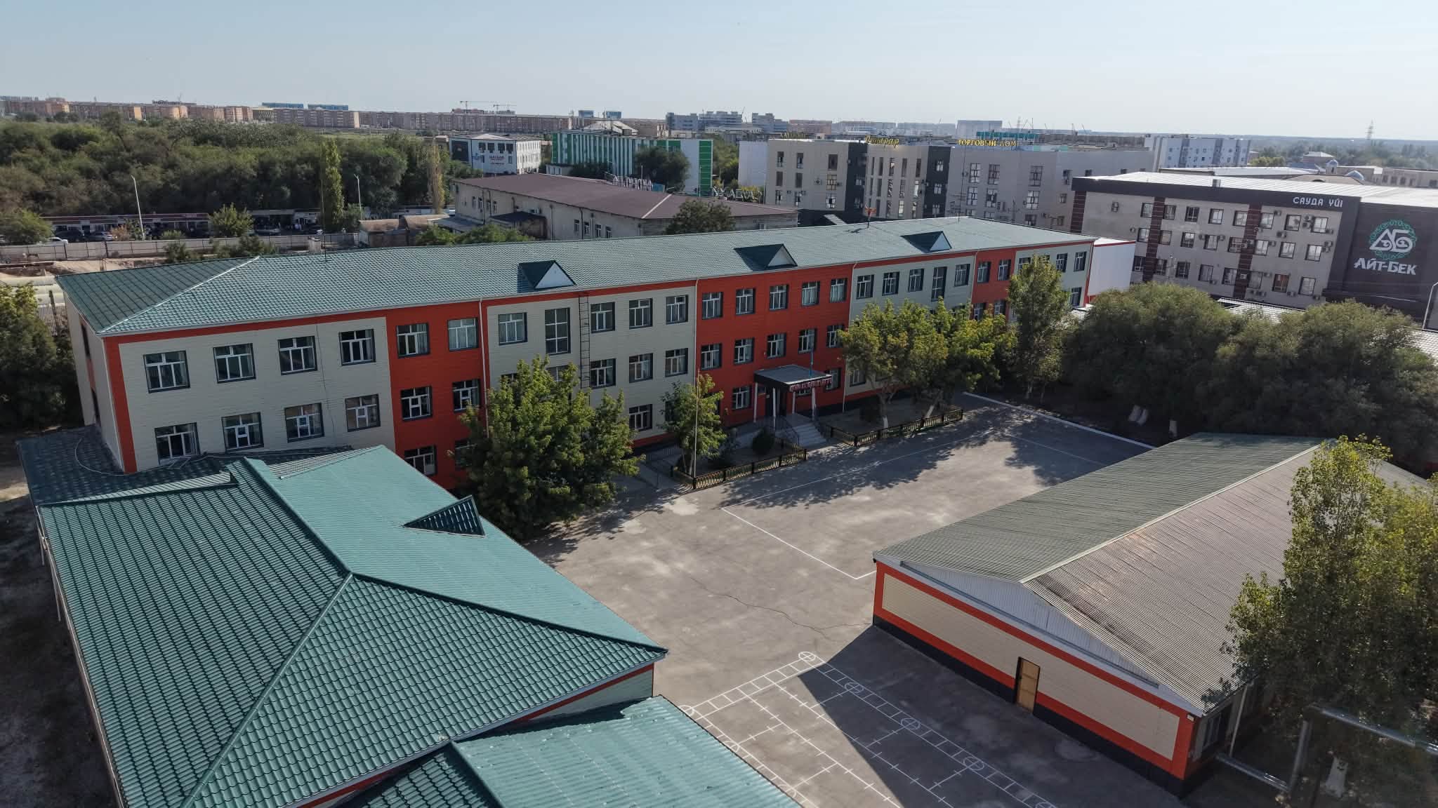 Жасыл шатырлы, қызғылт-сары қабырғалы мектеп ғимараты мен оның ауласының жоғарыдан көрінісі.