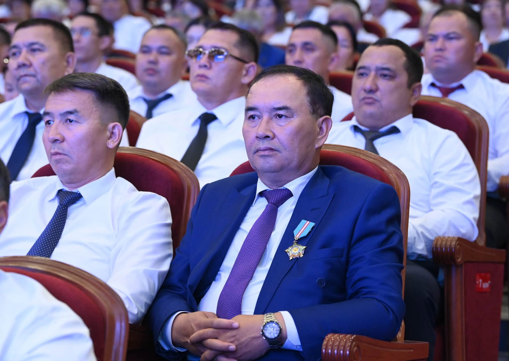 Ақ көйлек пен костюм киген ер адамдардың жиналыста отырғаны. Алдыңғы қатарда кеудесінде төсбелгісі бар ер адам.