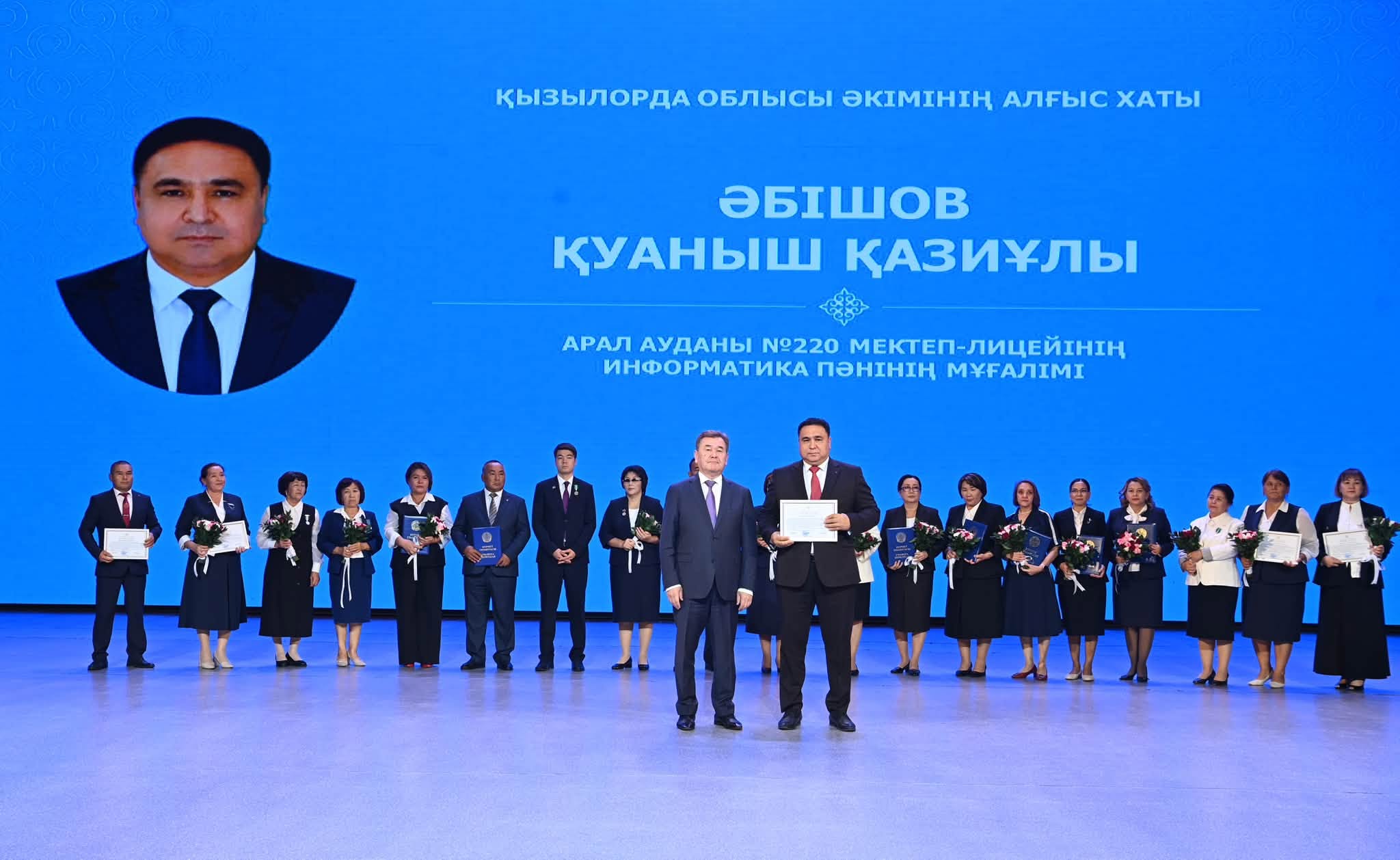 Марапатталған мұғалімдер тобы және артқы фондағы Әбішов Қуаныш Қазыұлының суреті мен алғыс хаты.