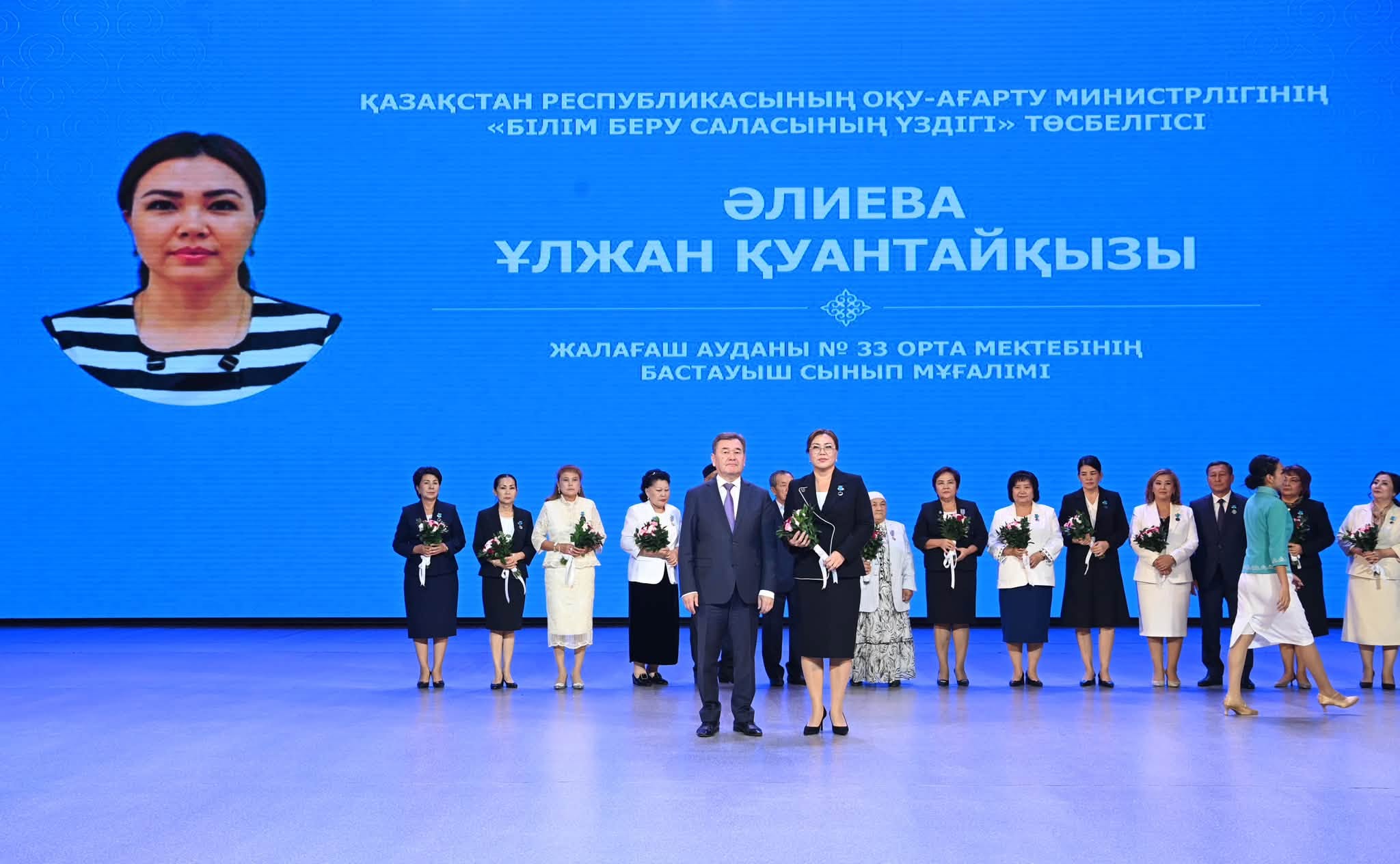Марапатталу салтанатындағы мұғалімдер мен Қазақстан Президенті. Фонда Әлиева Ұлжанның суреті мен есімі көрсетілген экран.