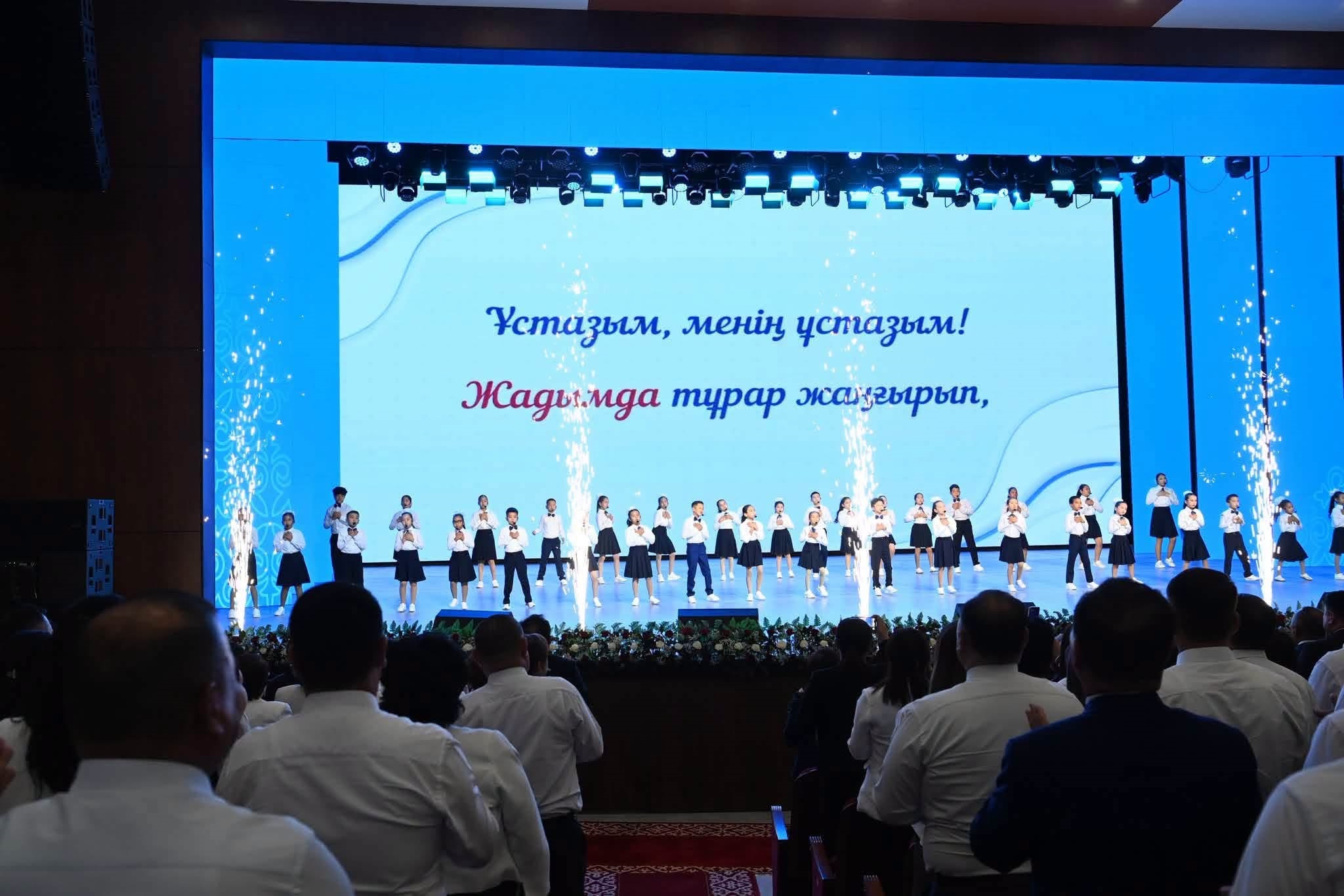 Сахнадағы балалардың қойылымы және артқы фондағы «Ұстазым, менің ұстазым!» деген жазуы бар экран.