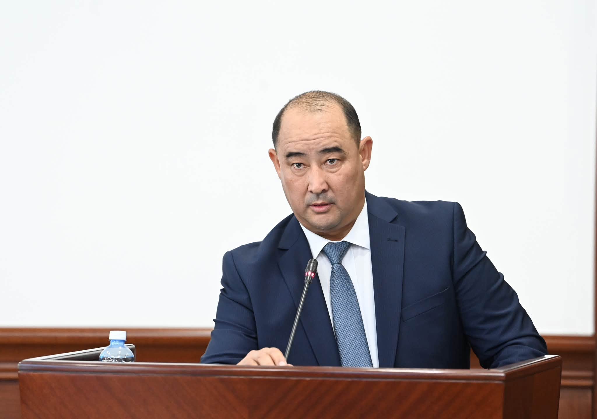 Көк костюм киген ер адам трибунада сөйлеп тұр.