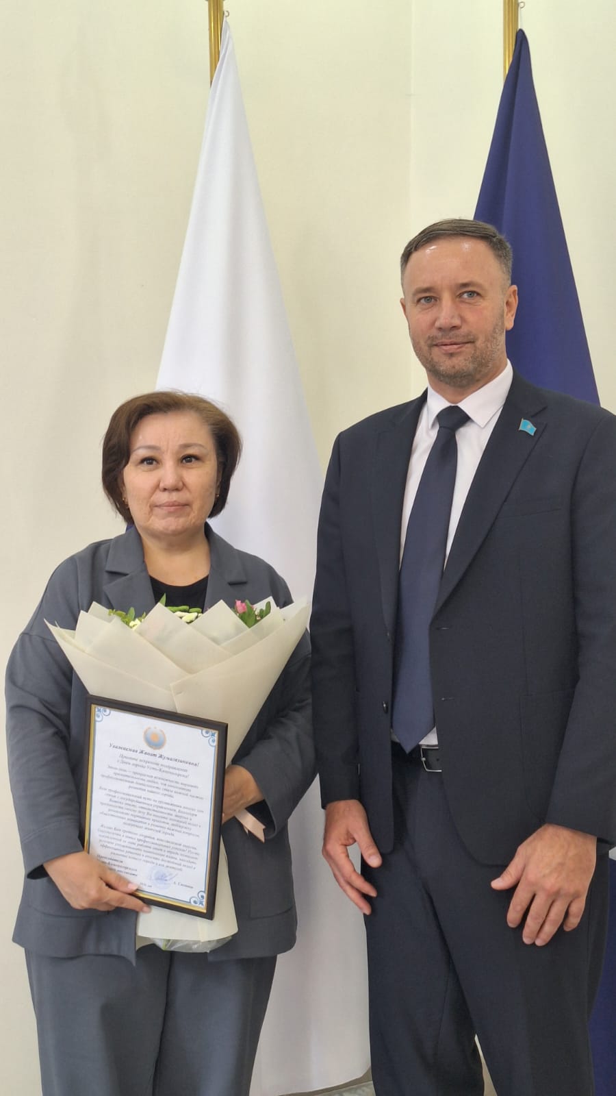 Гүл шоғы мен сауаттылығы бар әйел АҚ және көк жалаулардың фонында қара костюм киген адамның жанында тұрады.