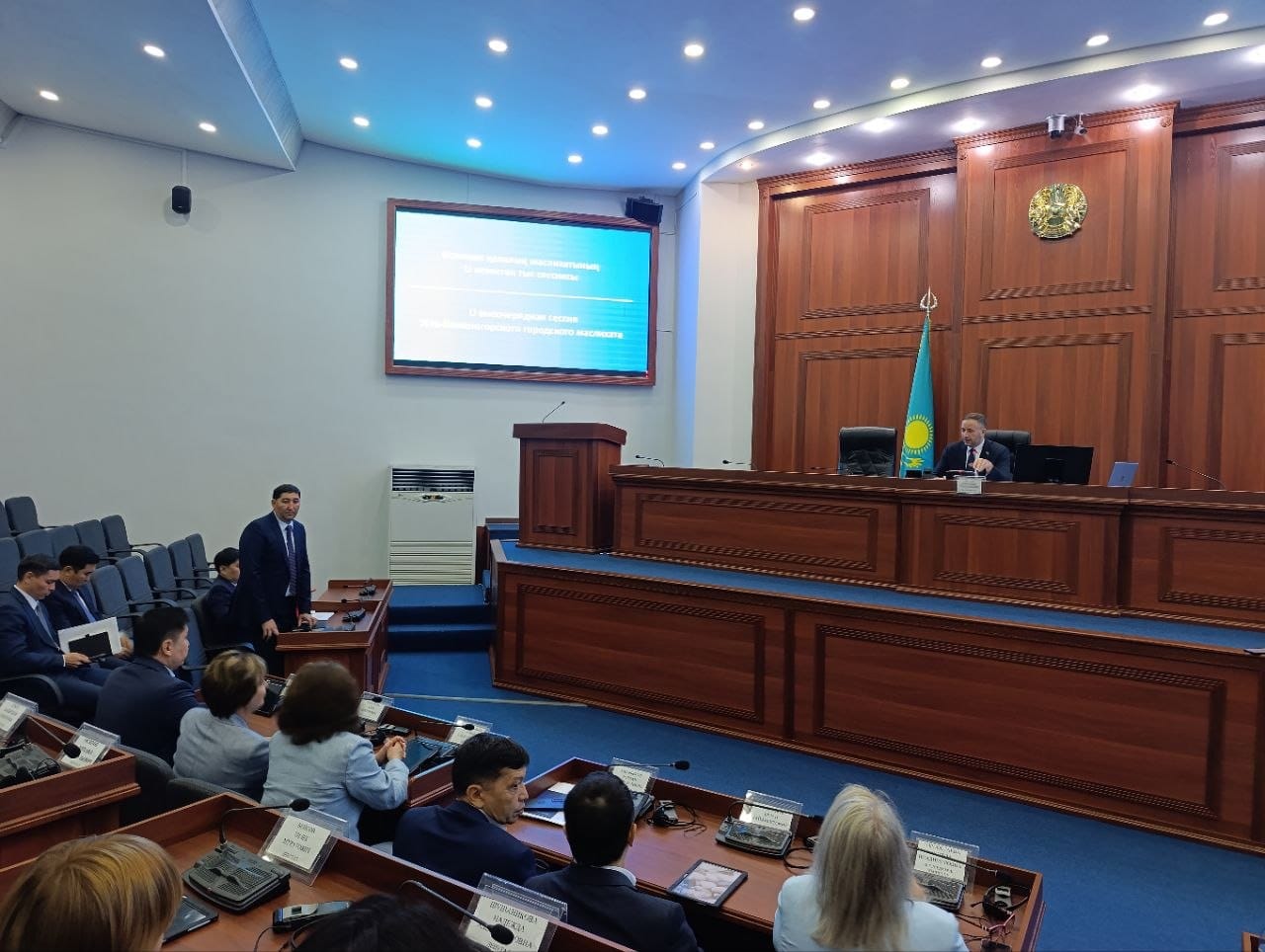 Конференц-залдағы отырыс: трибунаның артындағы спикер, аудитория және ағаш панельдердің фонында презентациясы бар экран.