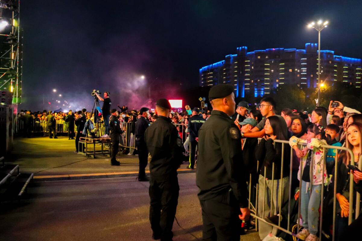 Полицейские следят за толпой людей за ограждением во время ночного мероприятия на фоне подсвеченного здания.