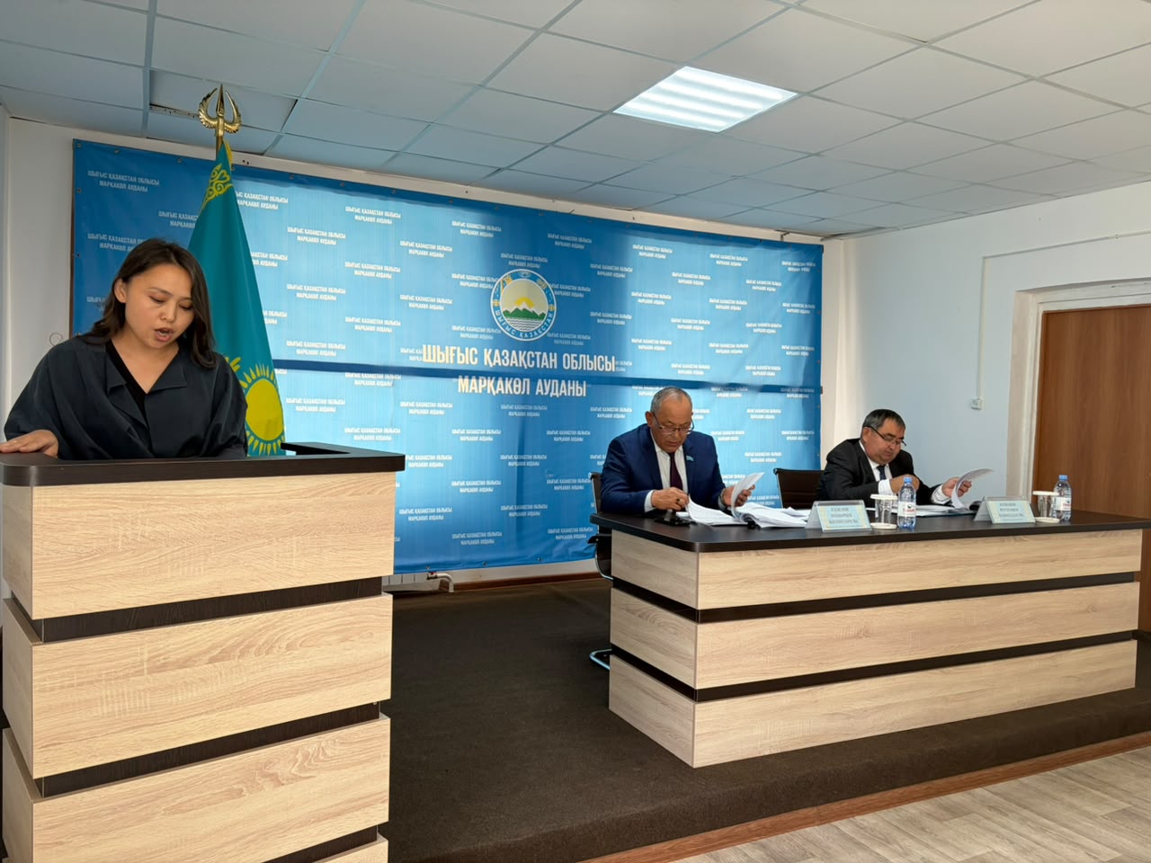Женщина выступает за трибуной перед двумя мужчинами на фоне синего баннера с гербом и надписью на казахском языке.
