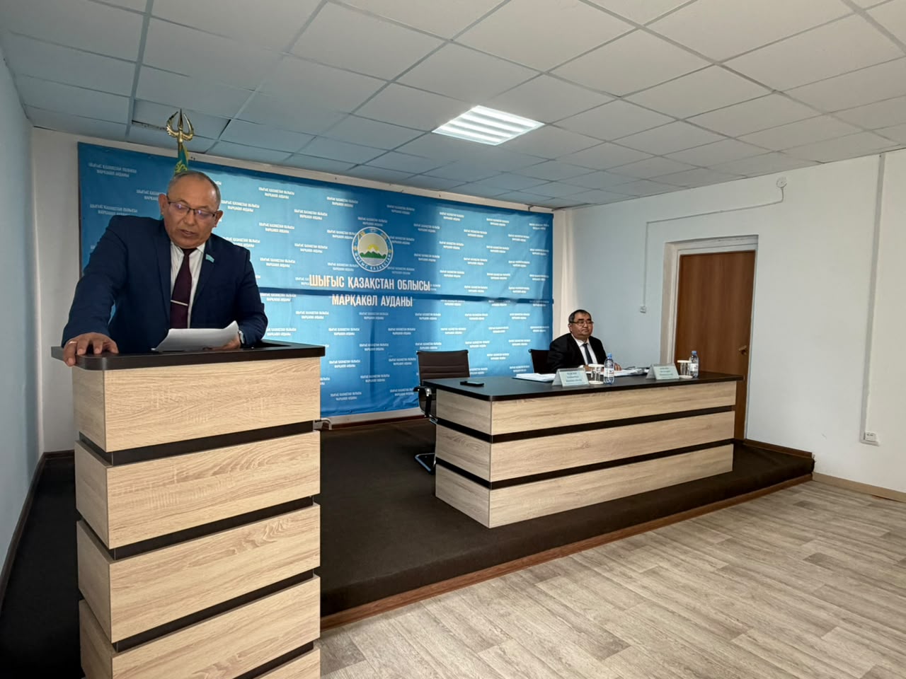 Мужчина в костюме выступает за трибуной на фоне синего баннера в официальном помещении.