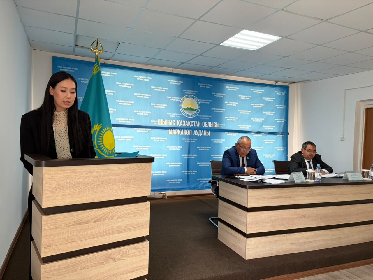 Женщина выступает за трибуной перед двумя мужчинами на фоне синего баннера с гербом и надписями на казахском языке.