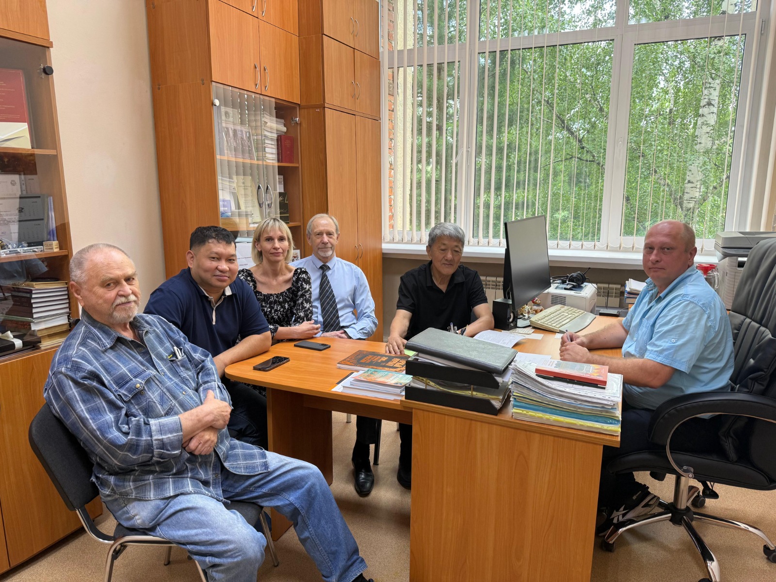 Группа из шести мужчин и одной женщины в офисе за рабочим столом на фоне книжных шкафов и окна.