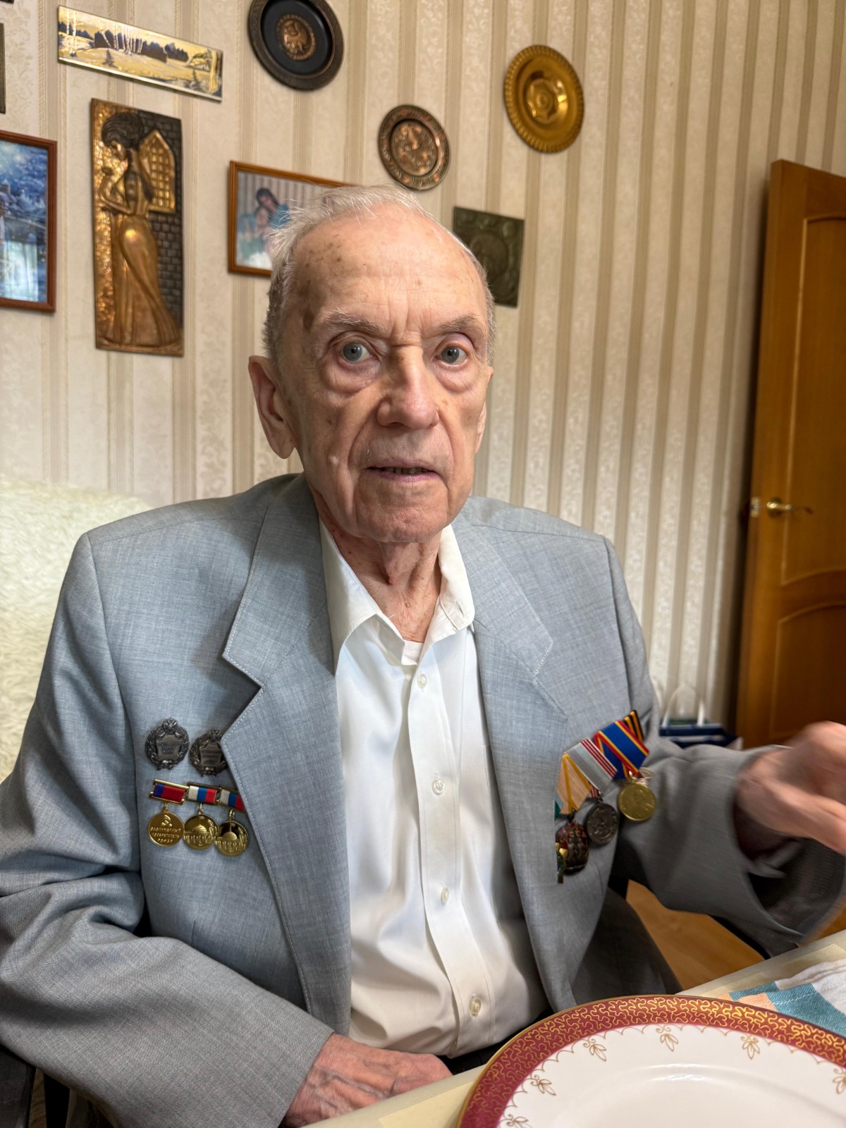 Доктор технических наук Олег Заякин в сером пиджаке с военными медалями и орденами на груди сидит за столом в комнате.