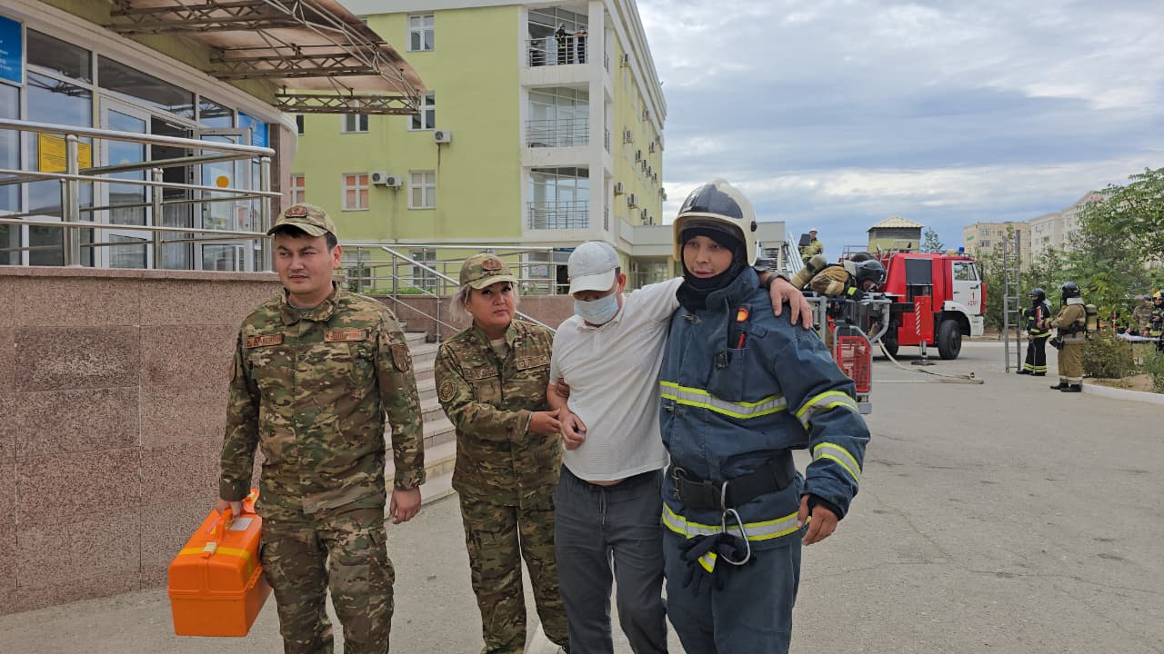 Құтқарушылар зардап шегушіні ғимарат алдынан көмектесіп шығарып жатыр.