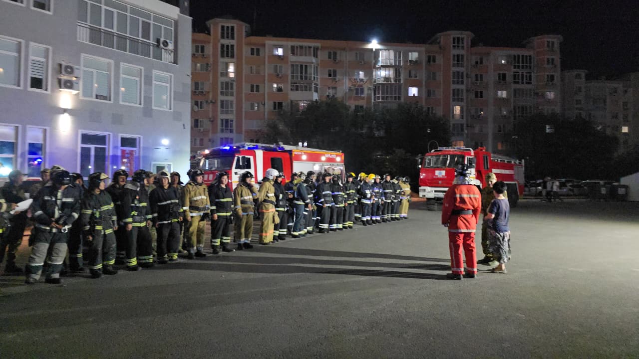 Түнгі уақытта өрт сөндіру машиналарының алдында сызылда тұрған өрт сөндірушілер тобы.