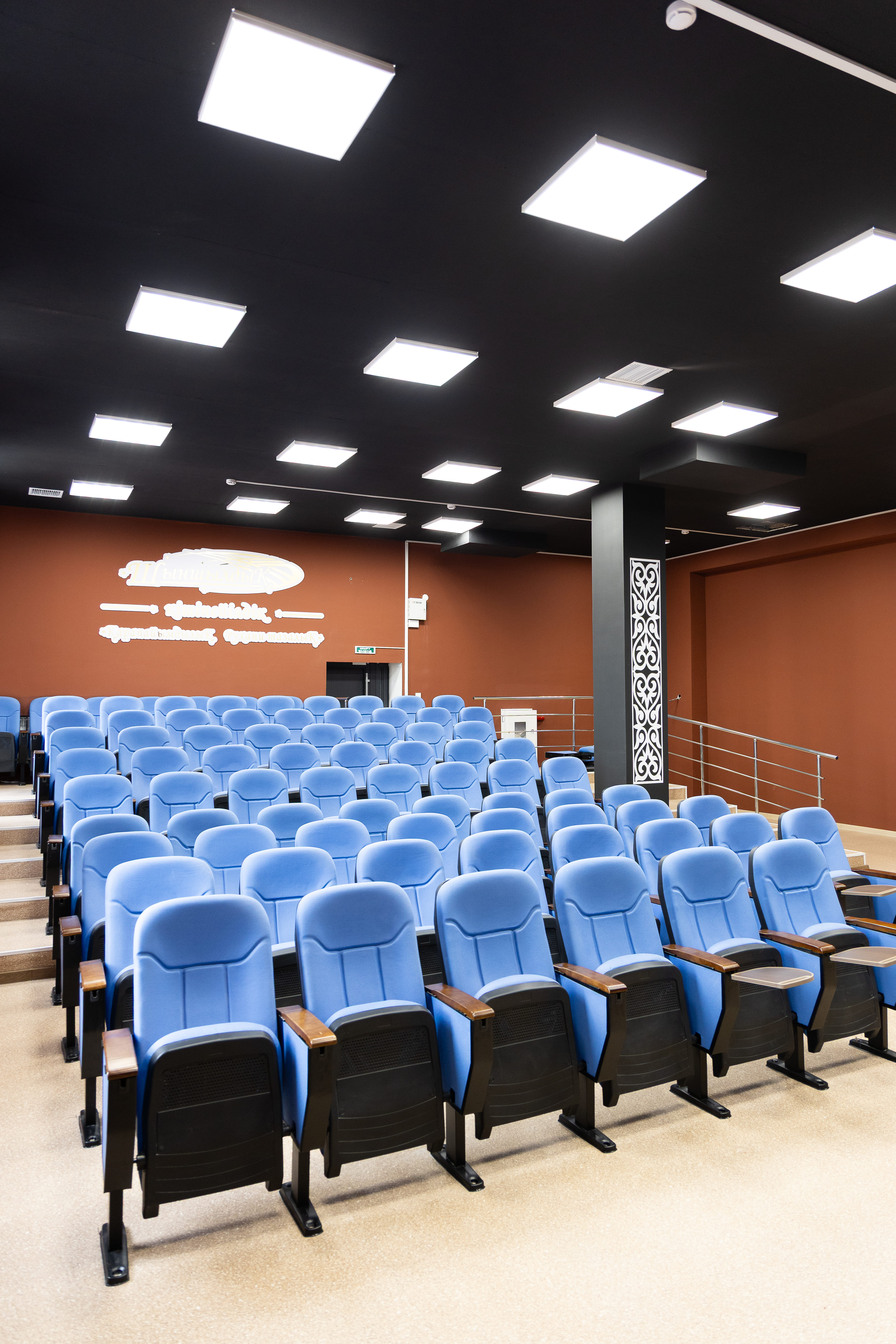 Ряды синих кресел в современном конференц-зале или кинотеатре с темным потолком и квадратными светильниками.