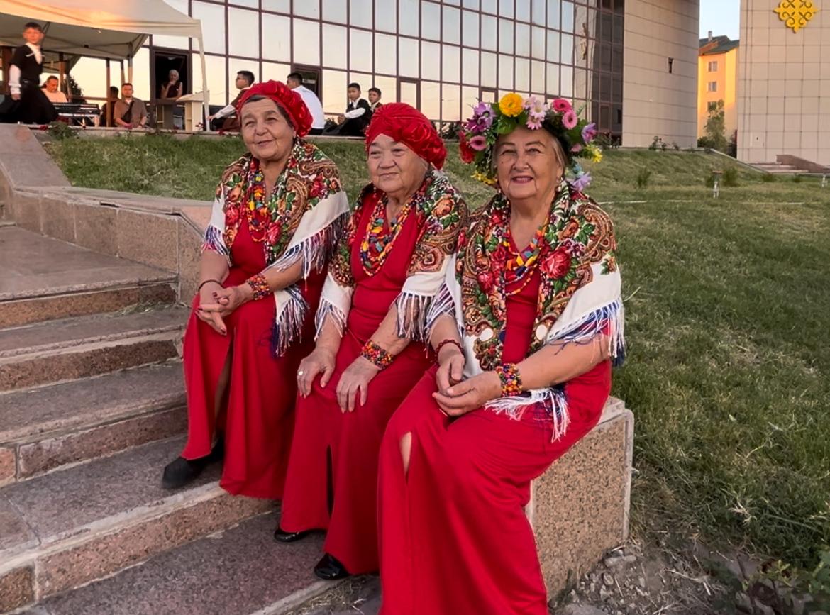 Ұлттық нақыштағы оюлы шәлі мен қызыл көйлек киген үш әйелдің сатыда отырған суреті.