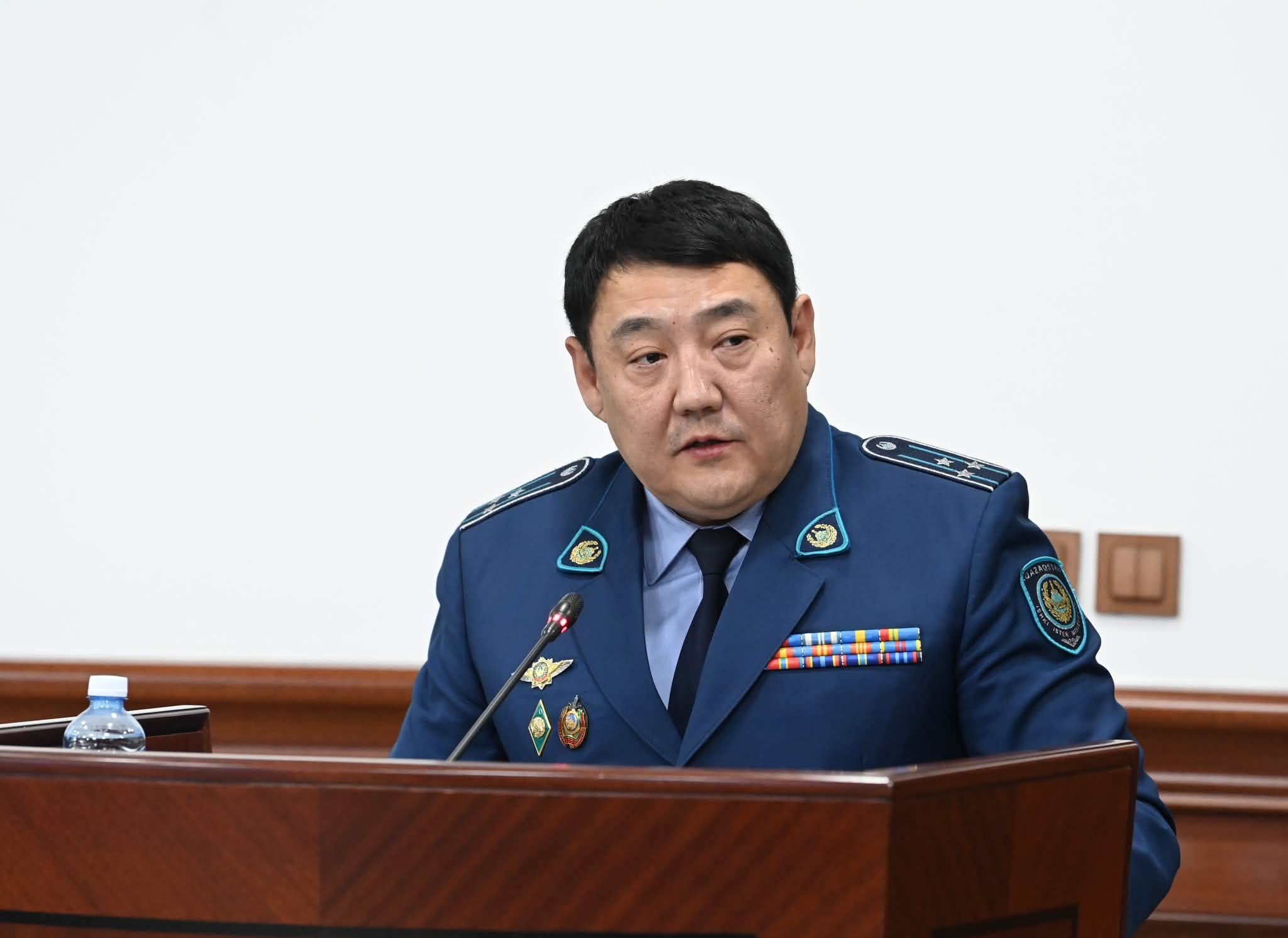 Көк форма киген, төсбелгілері бар ер адам трибунада сөйлеп тұр.