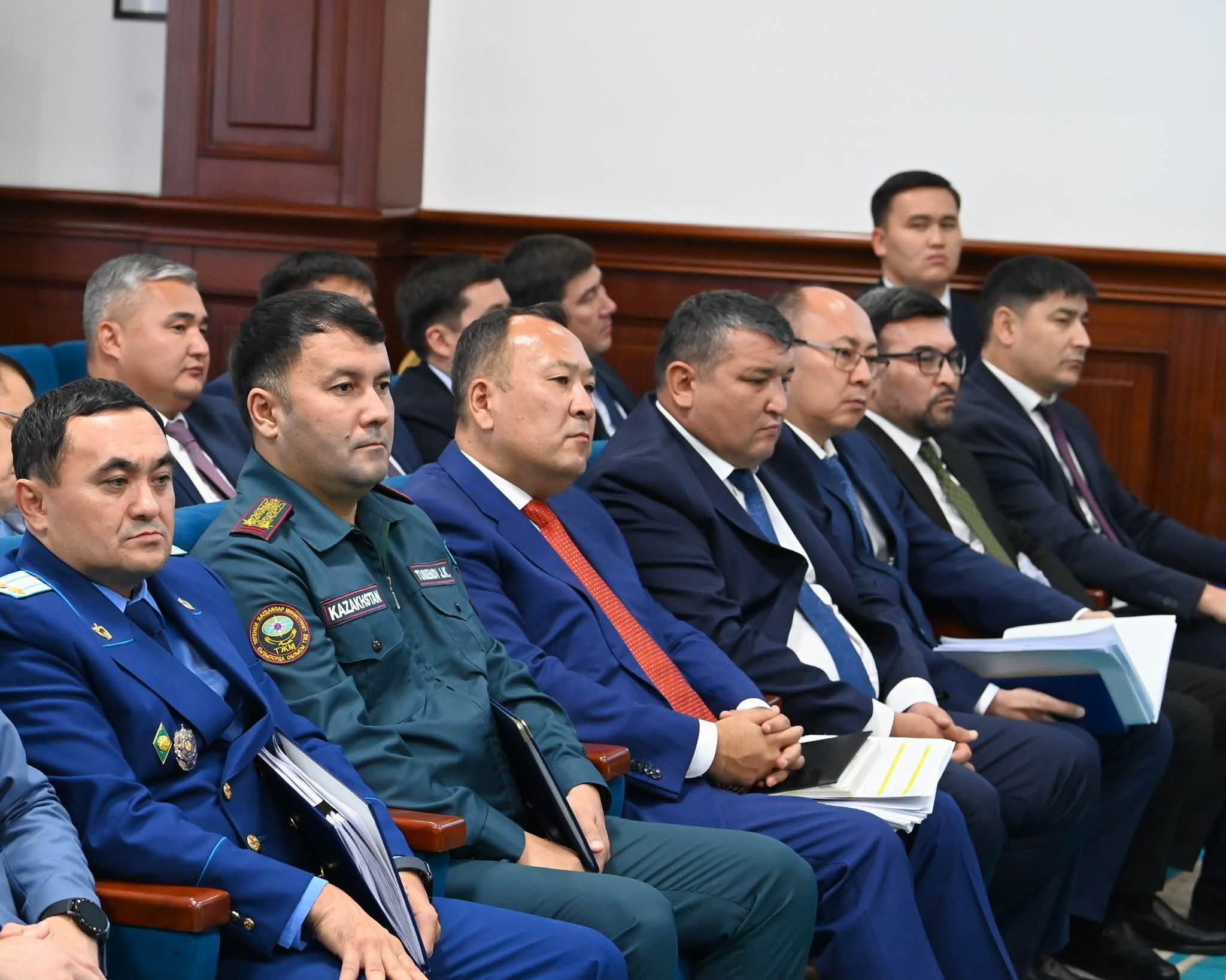 Жиналыста отырған ресми киімдегі және арнайы формадағы ер адамдар тобы.