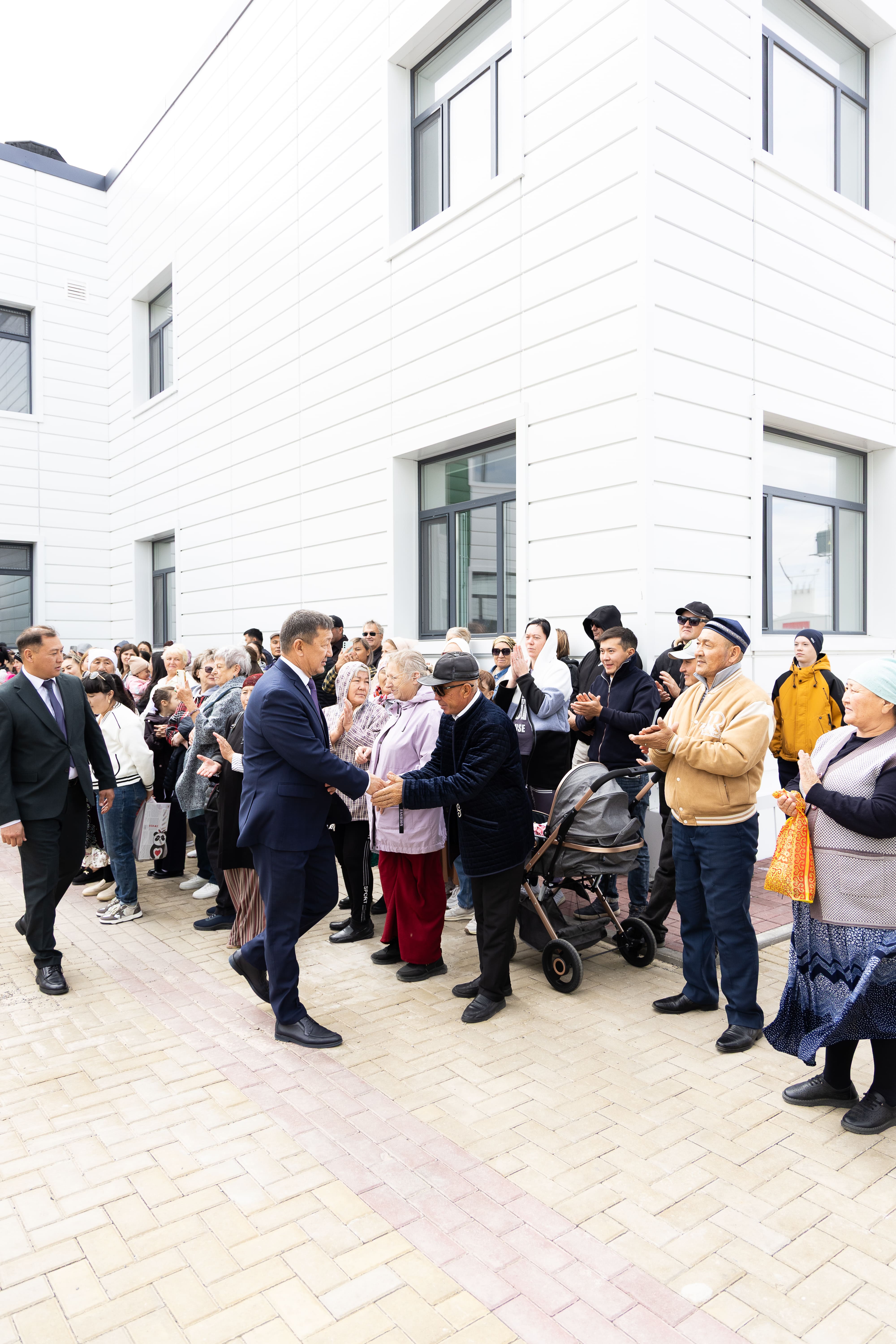 Мужчина в костюме пожимает руку пожилому человеку в окружении группы людей на фоне белого современного здания.