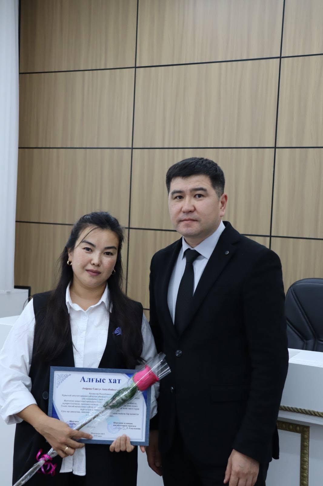 Алғыс хат пен гүл ұстаған әйел мен костюм киген ер адамның суреті.