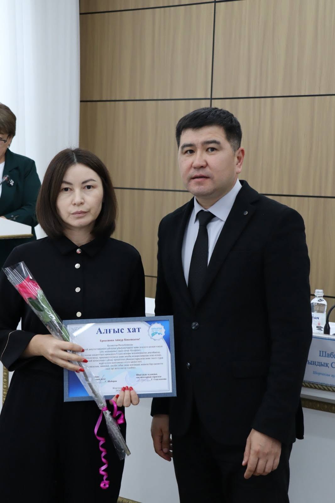Ресми киімдегі әйел мен ер адам. Әйел қолына алғыс хат пен гүл ұстап тұр.