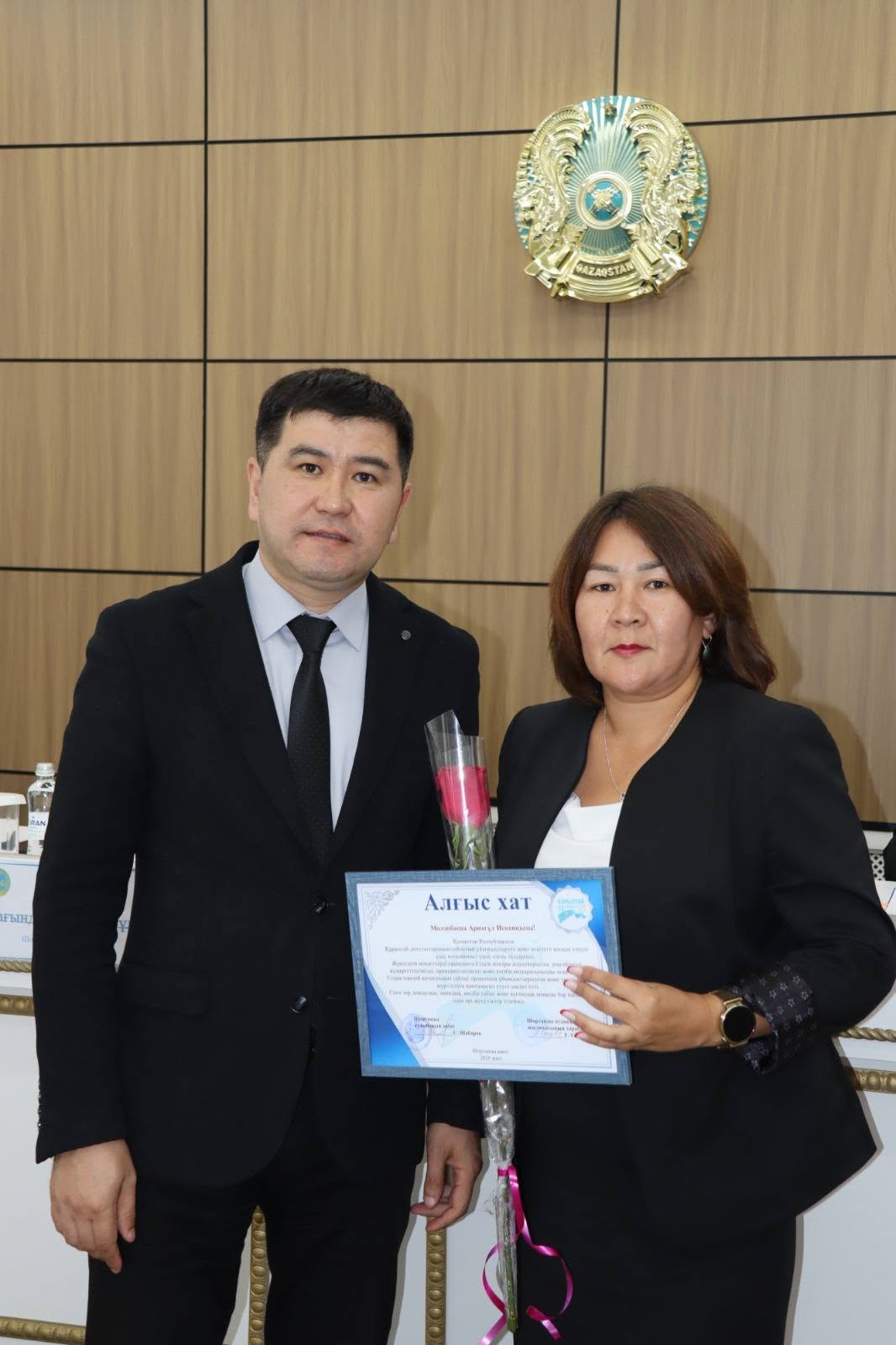 Ресми киімдегі ер адам мен әйел адамның алғыс хат пен гүл ұстап тұрған суреті.