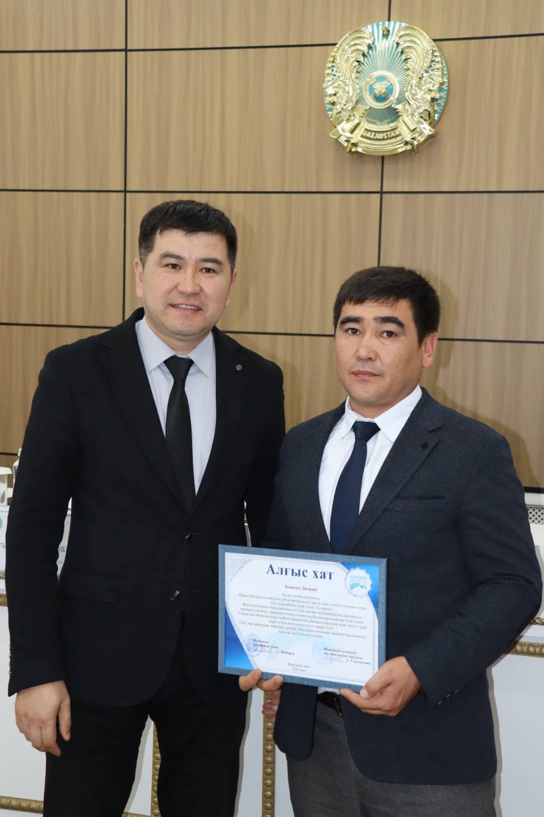 Екі ер адамның алғыс хатпен бірге түскен суреті. Фонда Қазақстан Республикасының гербі бейнеленген.