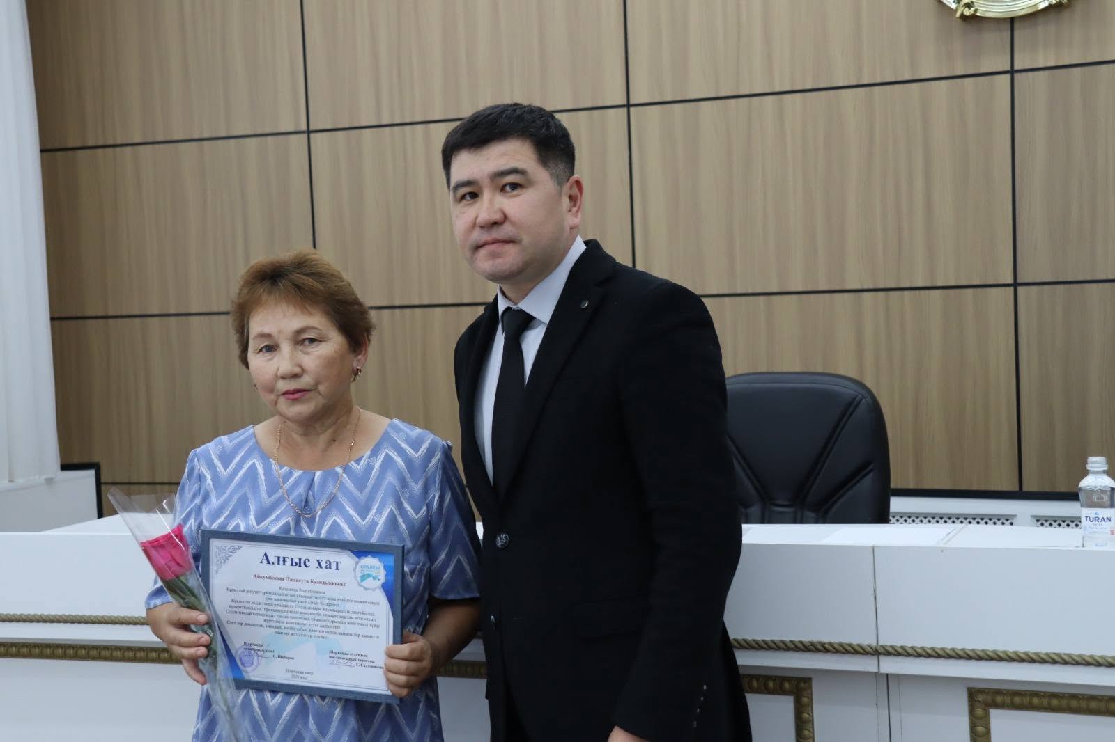 Алғыс хат пен гүл ұстаған әйел және қасында тұрған костюм киген ер адам.