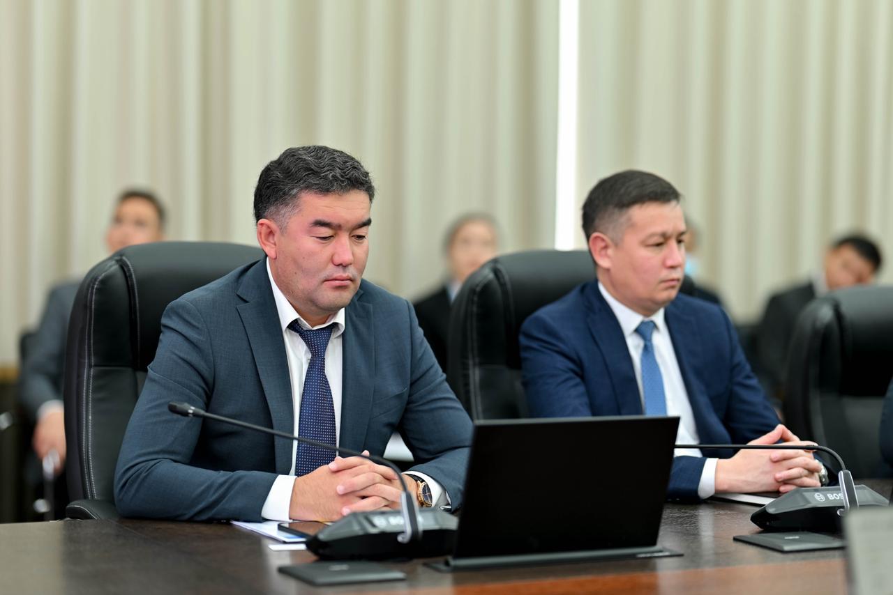 Жиналыста ноутбук пен микрофон алдында отырған костюм киген екі ер адам.