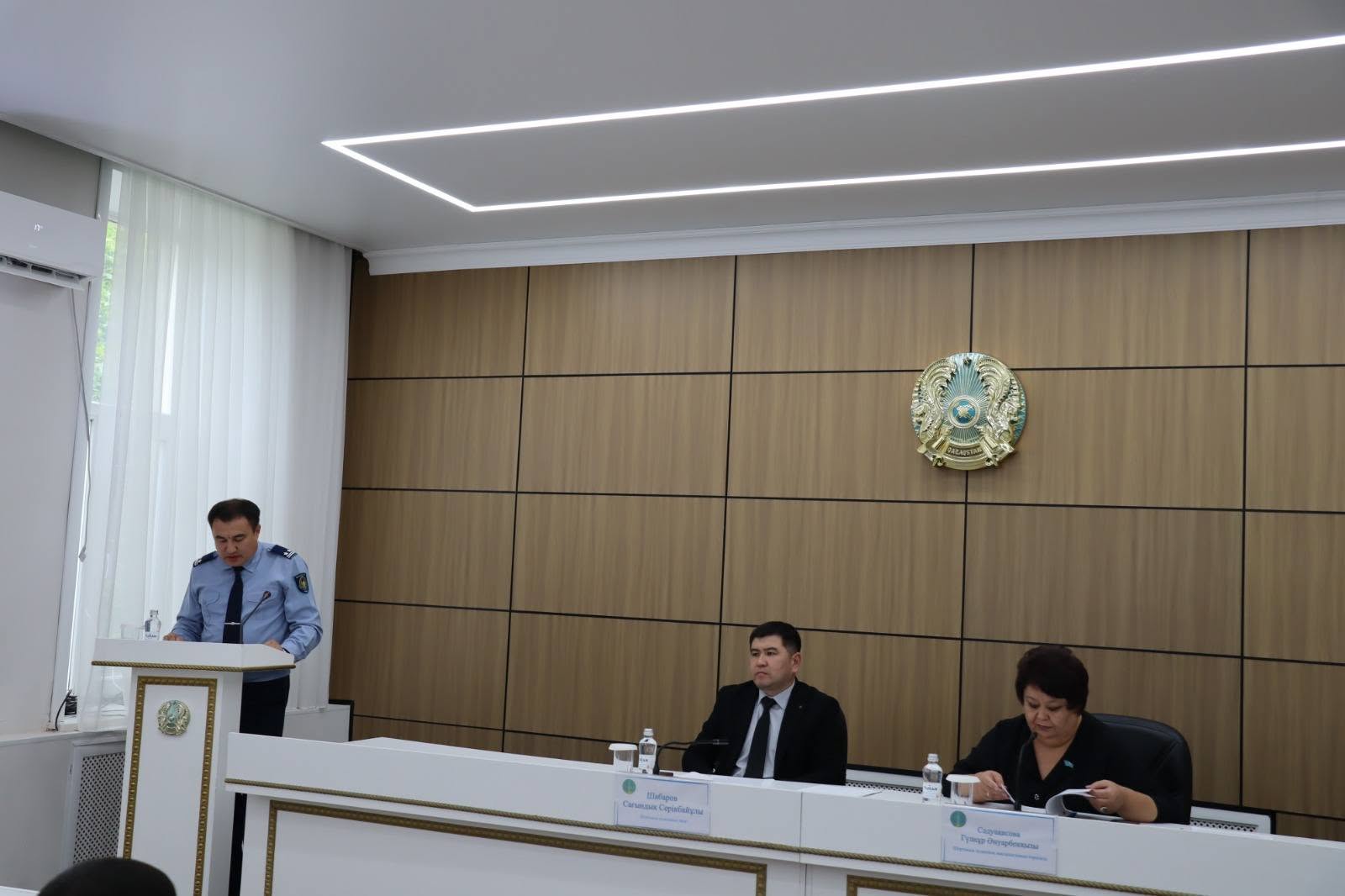 Ресми жиналыста сөйлеп тұрған полиция қызметкері және үстелде отырған тыңдаушылар.
