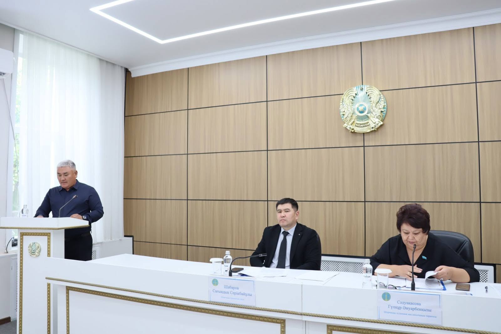 Ресми жиналыста сөйлеп тұрған спикер және үстел басында отырған комиссия мүшелері.