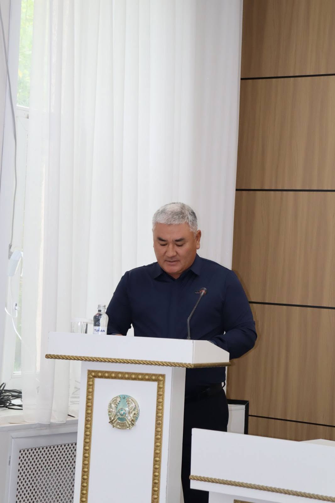 Трибунада сөйлеп тұрған қара көйлек киген ер адам.
