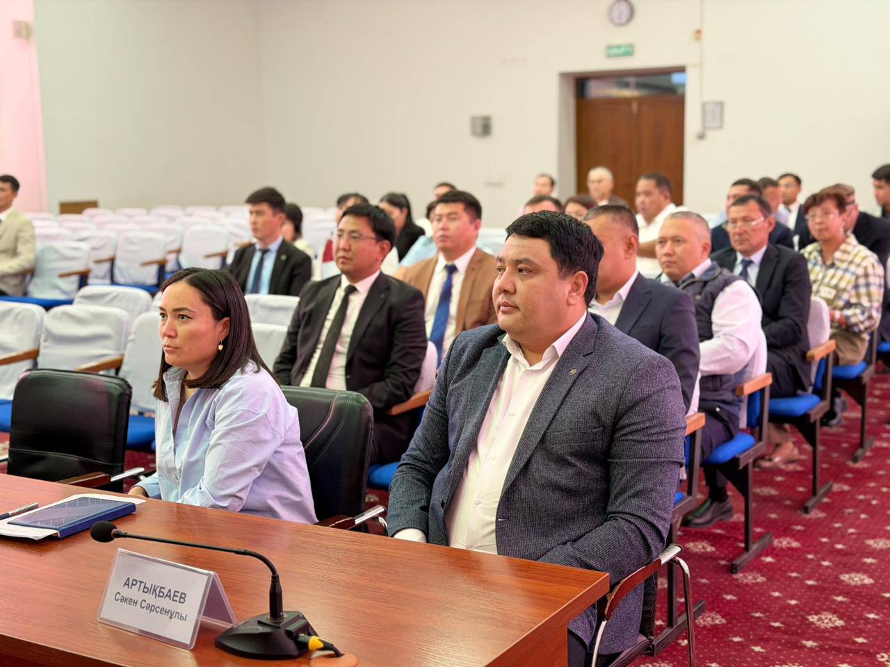 Жиналысқа қатысып отырған ресми киінген адамдар тобы.