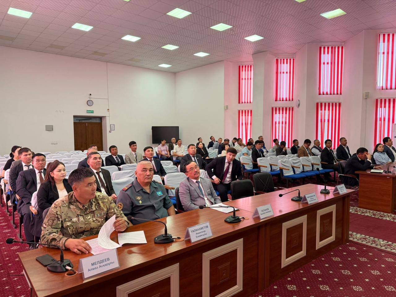 Конференция залында жиналған ресми киімдегі және әскери формадағы адамдар.