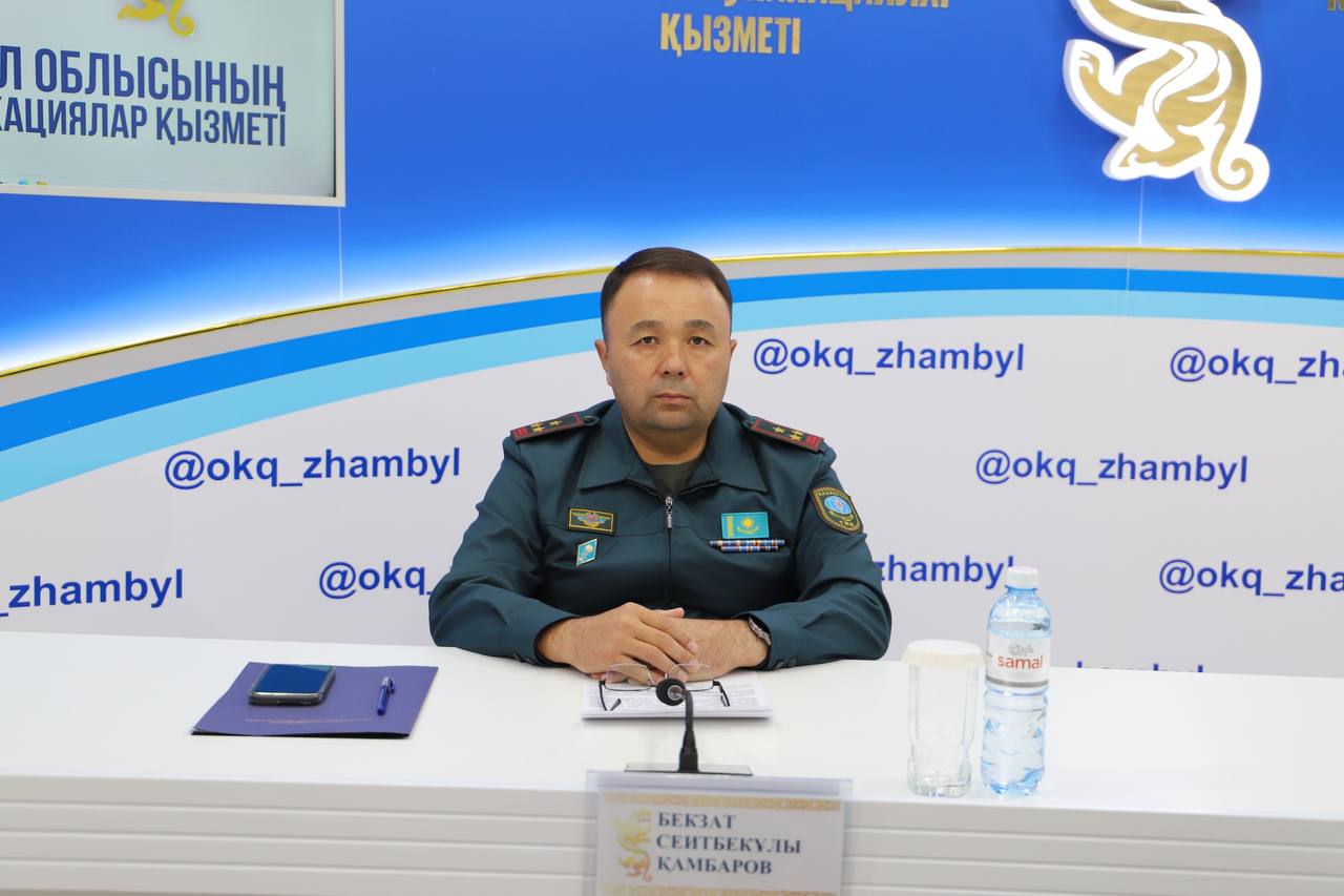 Әскери формадағы ер адам үстелде отыр. Алдында есімі жазылған тақта мен су бутылкасы бар.
