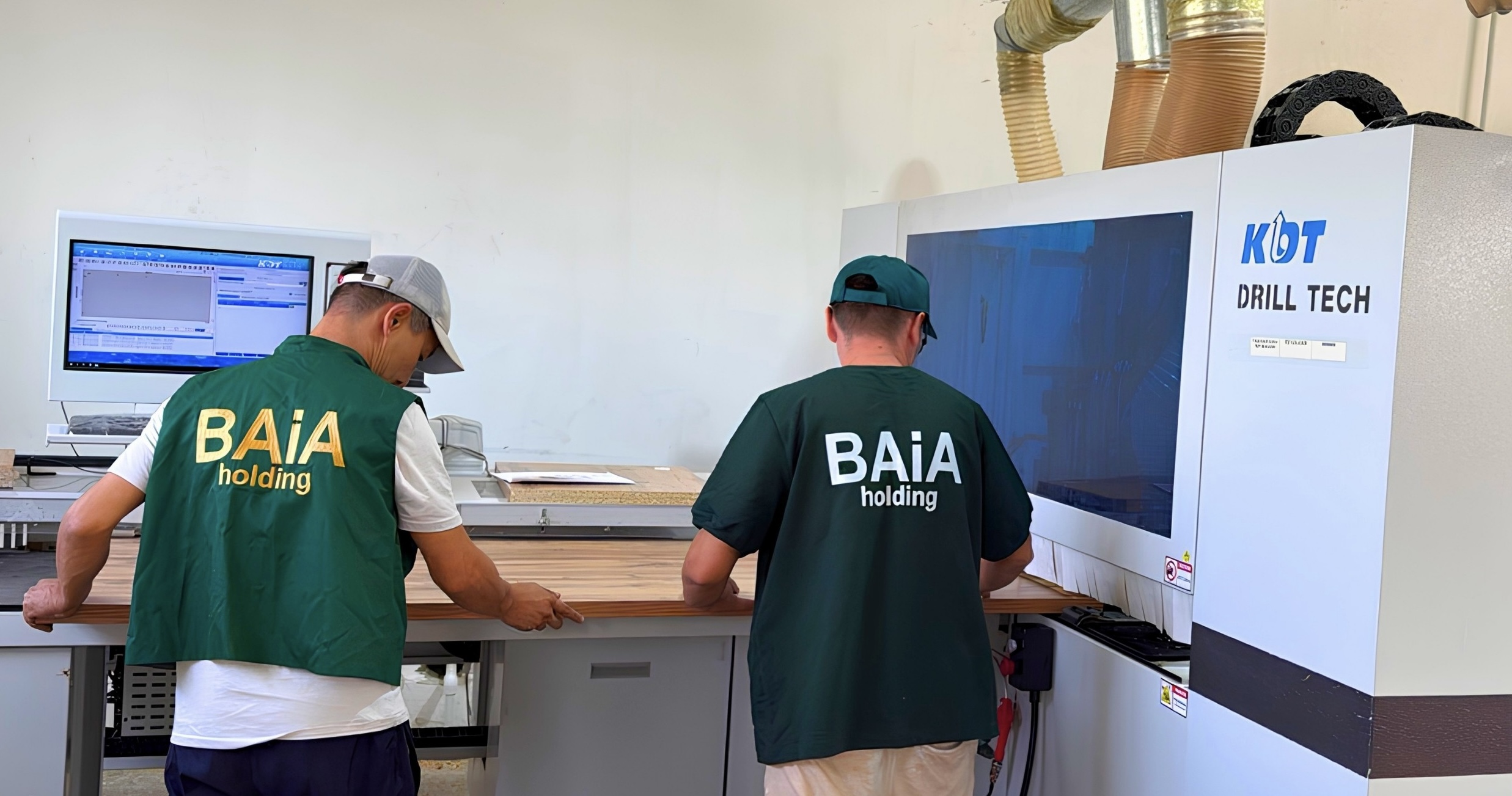 Двое рабочих в форме BAIA holding работают с оборудованием KDT Drill Tech в цеху.