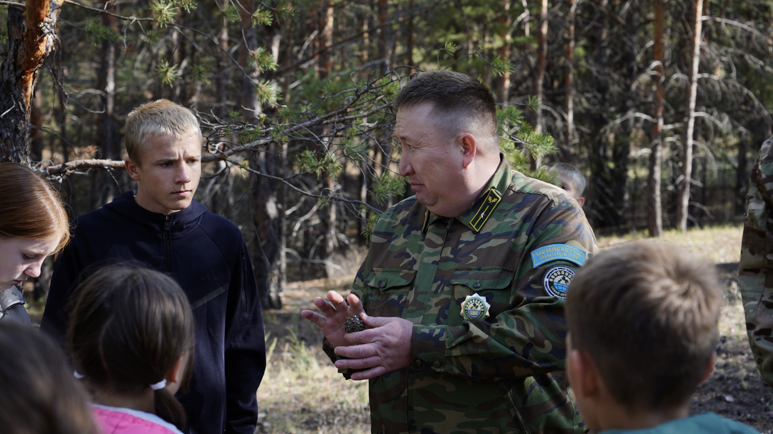 Әскери формадағы ер адам орман ішінде балаларға бір нәрсені түсіндіріп жатыр.
