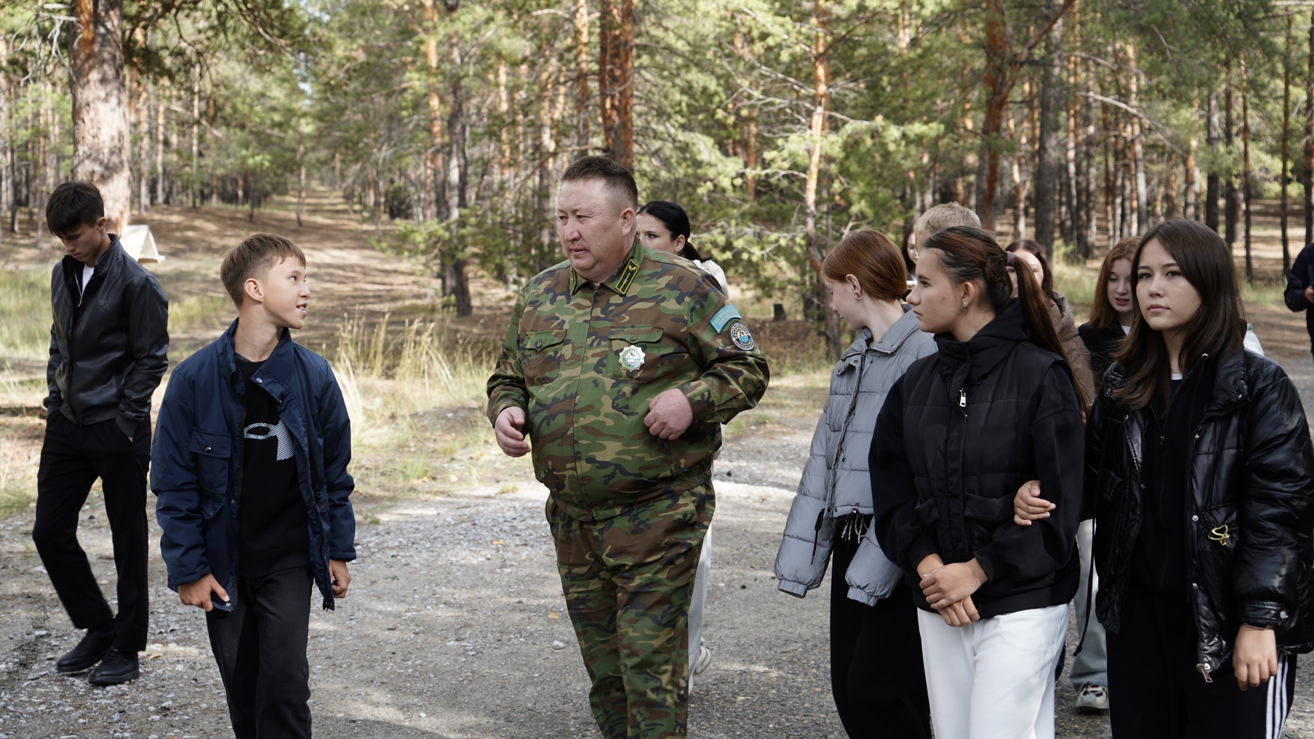 Орманды жерде әскери формадағы адамның жасөспірімдер тобымен сөйлесіп, жүріп бара жатқаны.