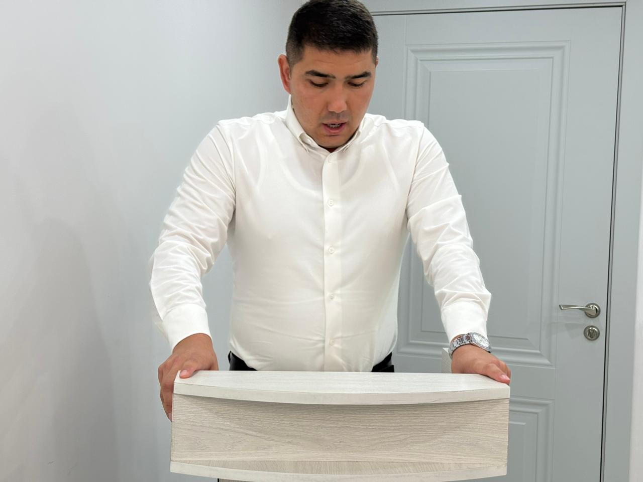 Ақ жейде киген ер адам ағаштан жасалған жиһазды ұстап тұр.
