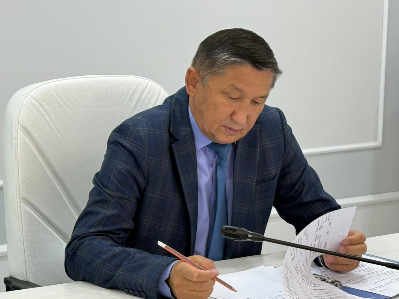 Көк костюм киген ер адам үстел басында құжаттарды қарап, қағазға жазып отыр.