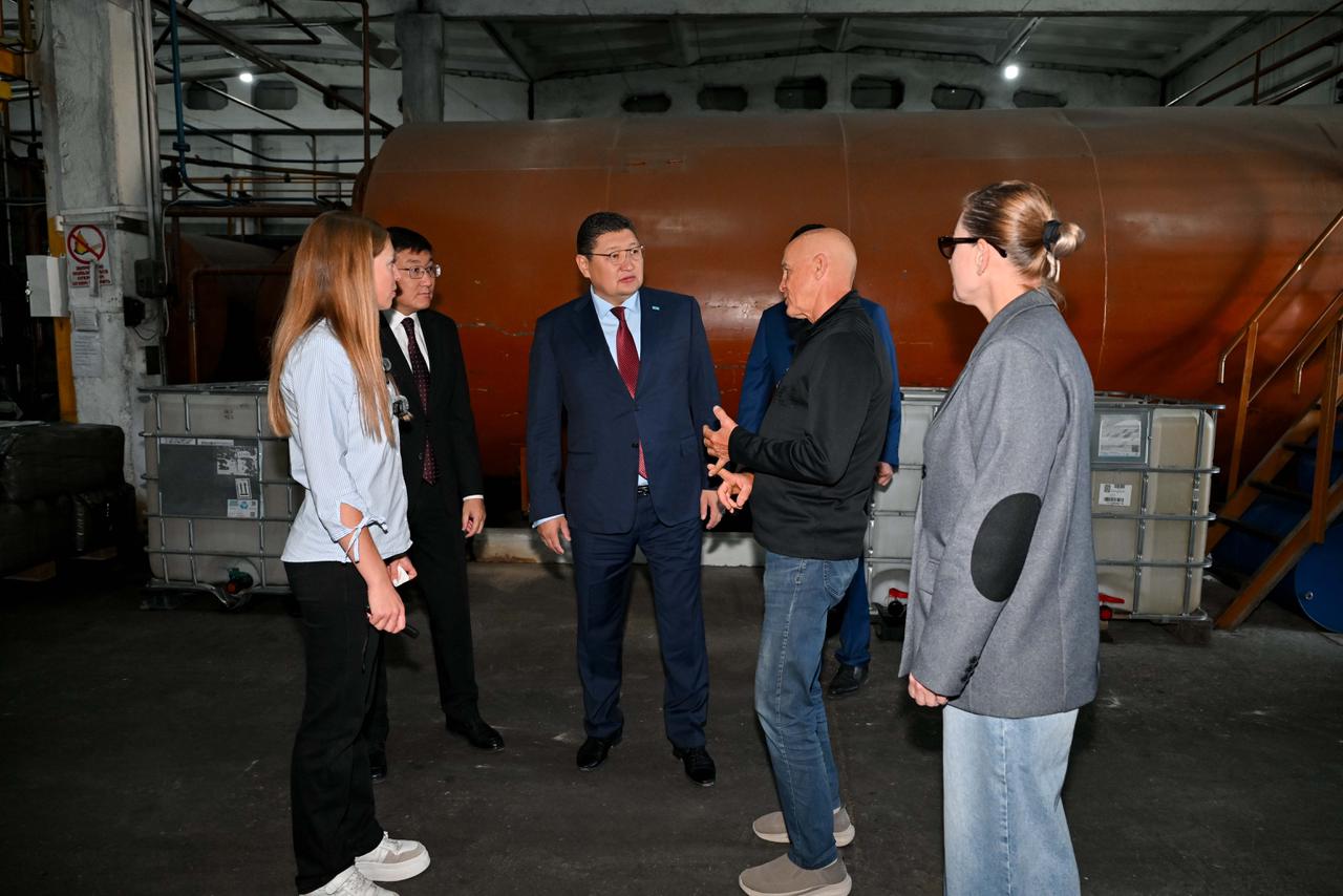 Өндіріс цехындағы үлкен резервуардың жанында тұрып, әңгімелесіп жатқан адамдар тобы.