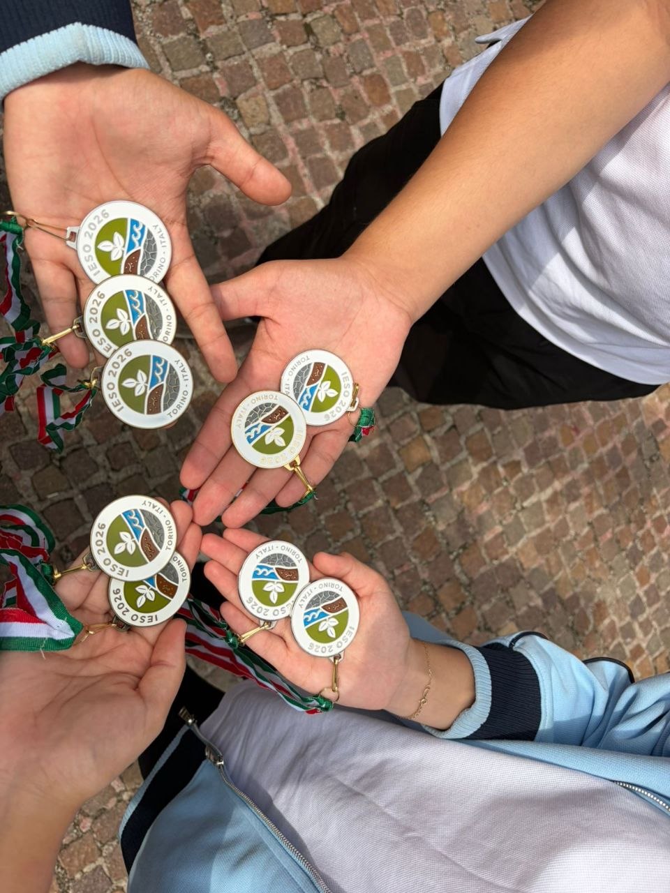 Несколько рук держат памятные медали с надписью «Torino Italy IESO 2026» на фоне брусчатки.