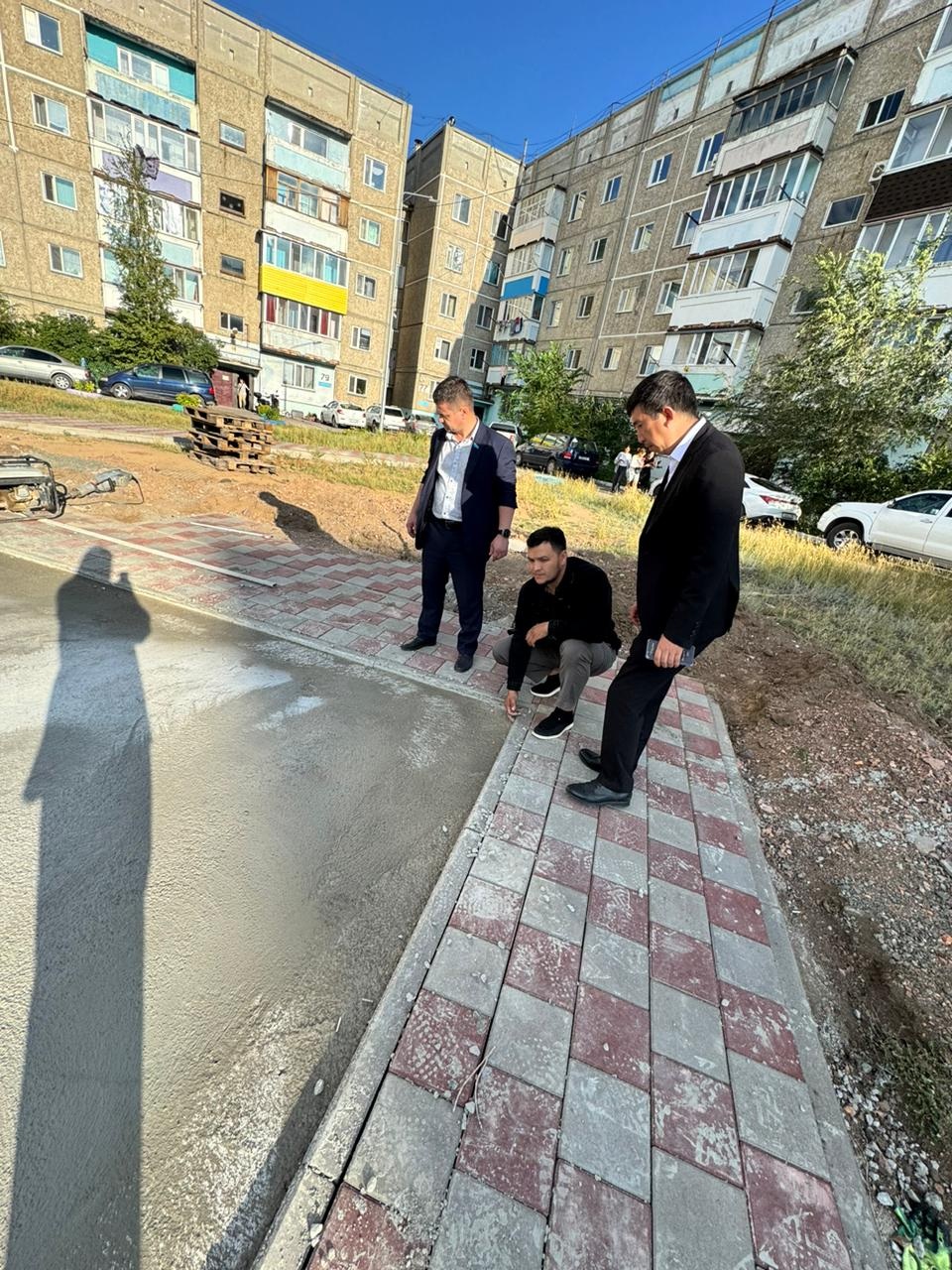 Көпқабатты үйлердің алдындағы жалау жолдың жағдайын тексеріп жатқан үш ер адам.
