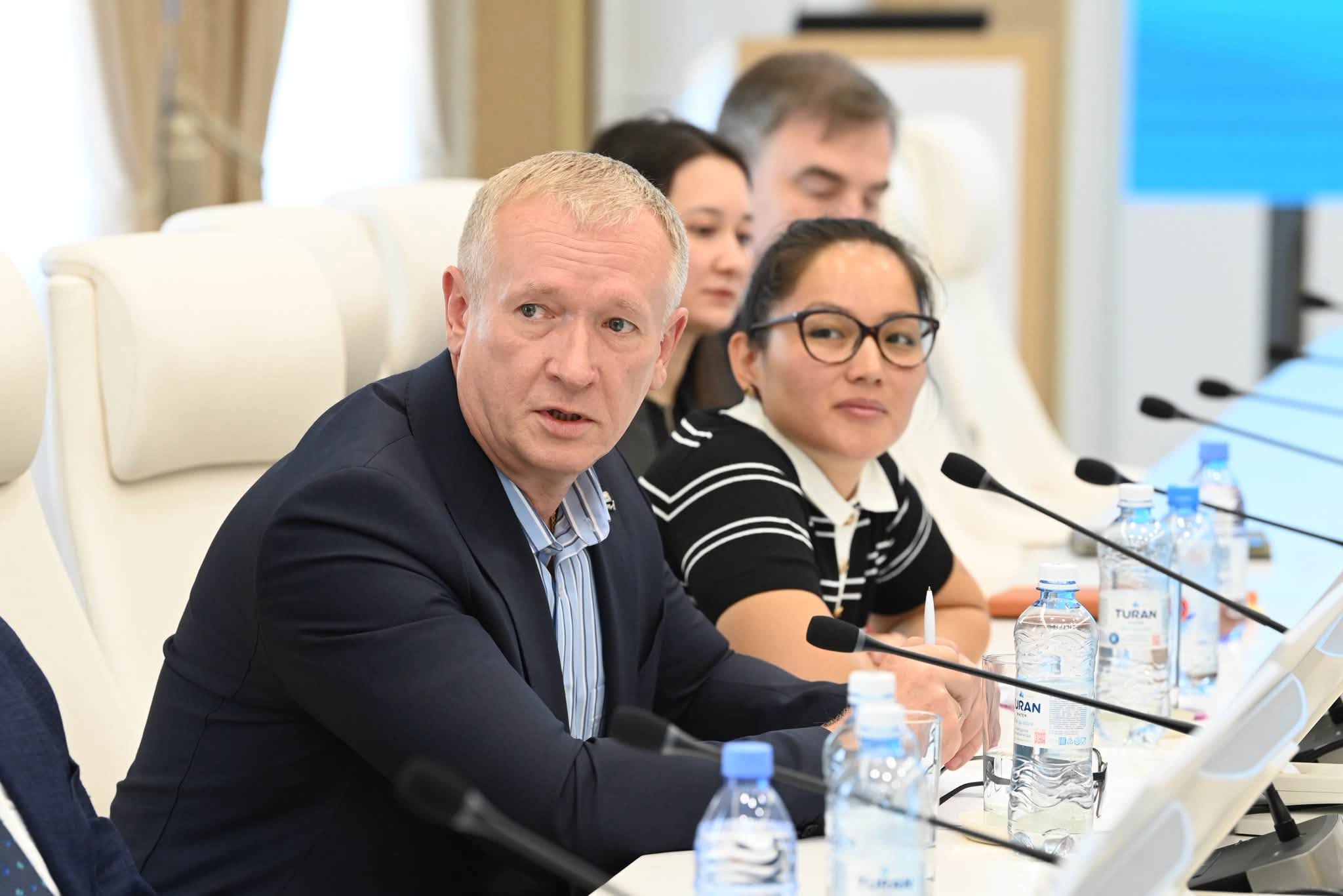 Жиналыста сөйлеп жатқан костюм киген ер адам және оның қасында отырған әріптестері.